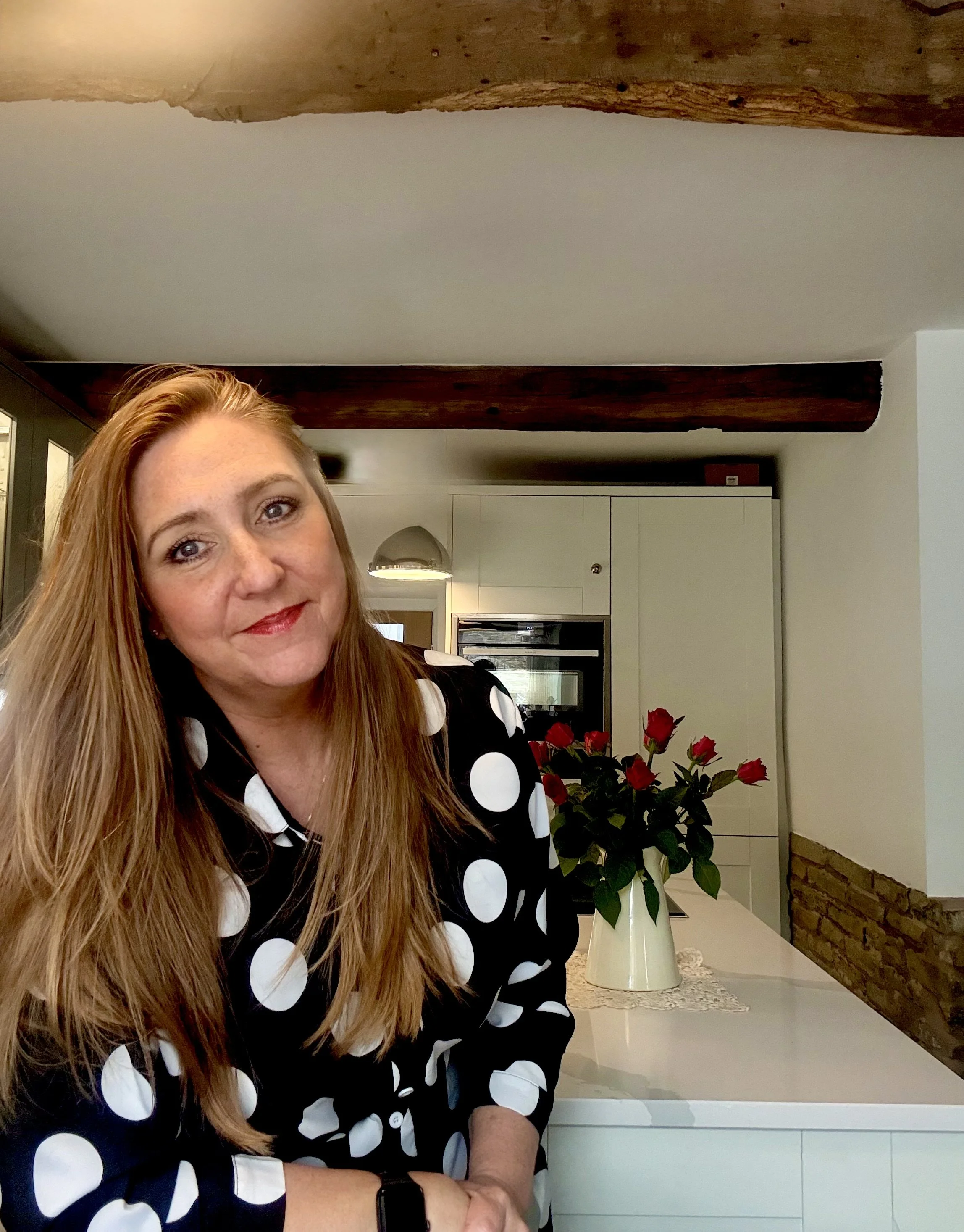 Katie Flanagan, Ribble Valley, Lake District and Yorkshire Dales Independent Wedding Celebrant, standing in her Yorkshire Dales kitchen, she’s wearing a black dress with large white polka dots, there’s a bouquet of red roses on the counter.