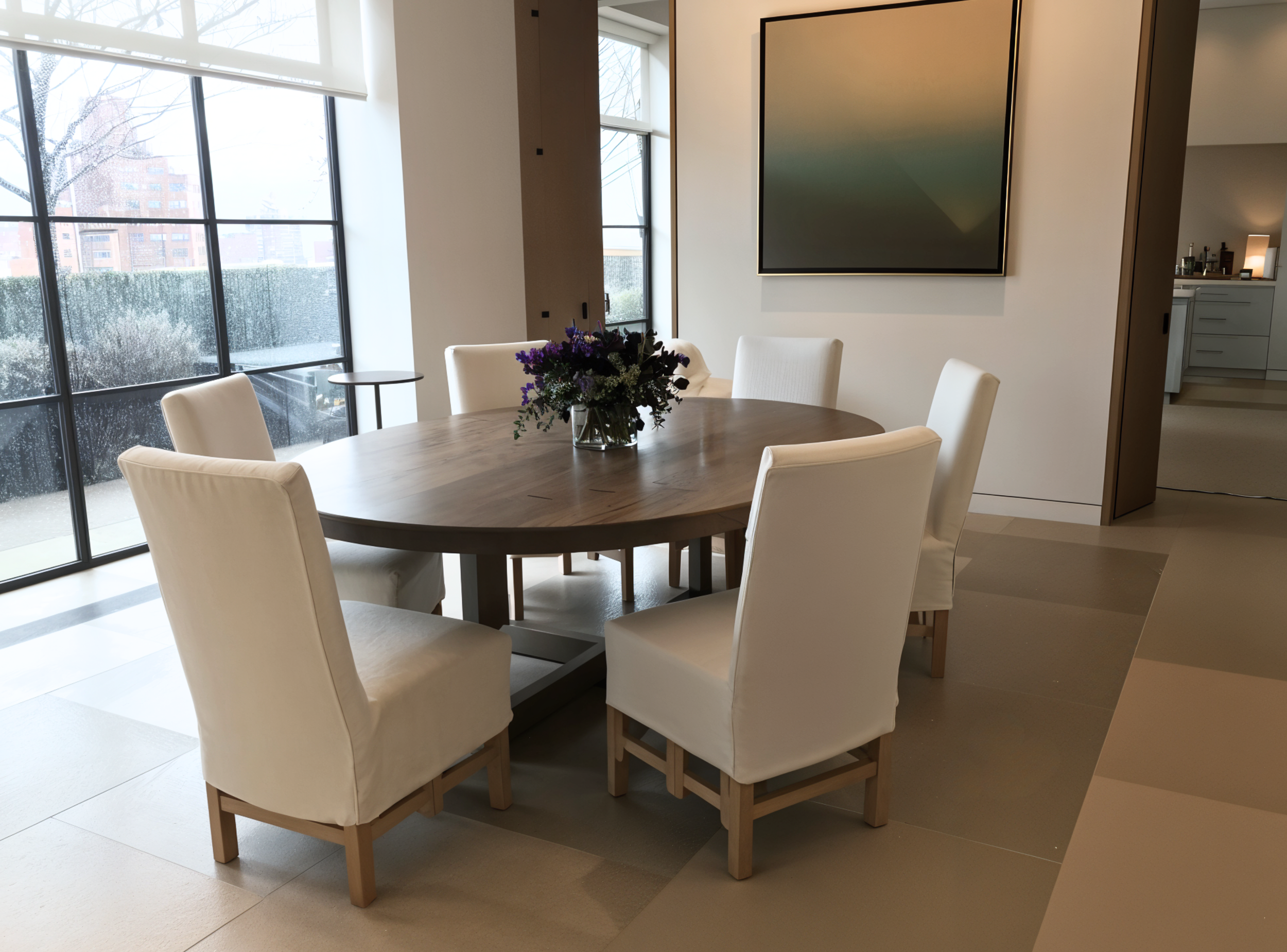 Modern dining room with a round wooden table, surrounded by six white upholstered chairs. A vase of purple flowers sits on the table. Large windows and contemporary artwork are visible in the background.