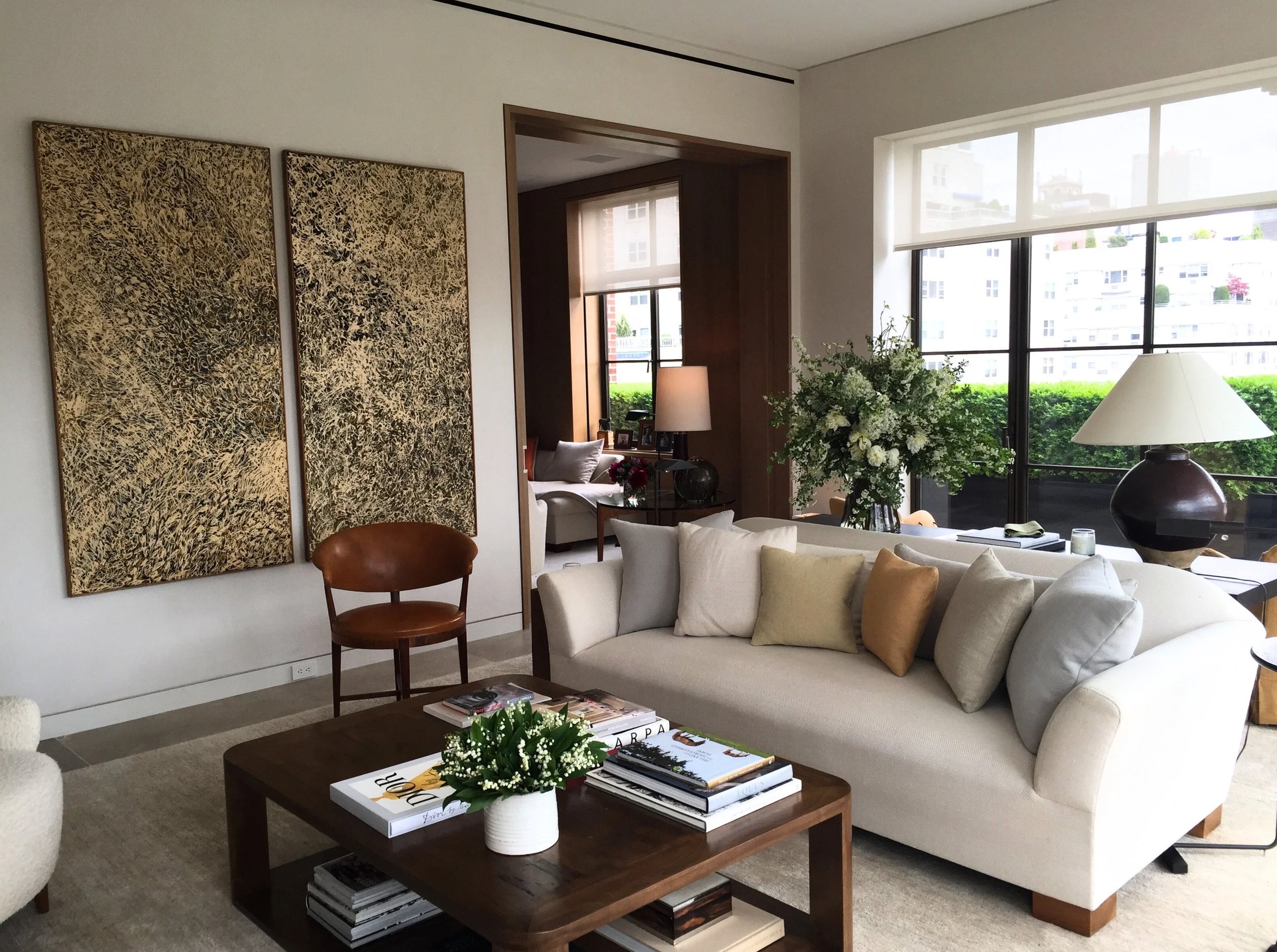 Modern living room with beige sofa, colorful pillows, wooden coffee table with books, large abstract art pieces on the wall, and potted plants.