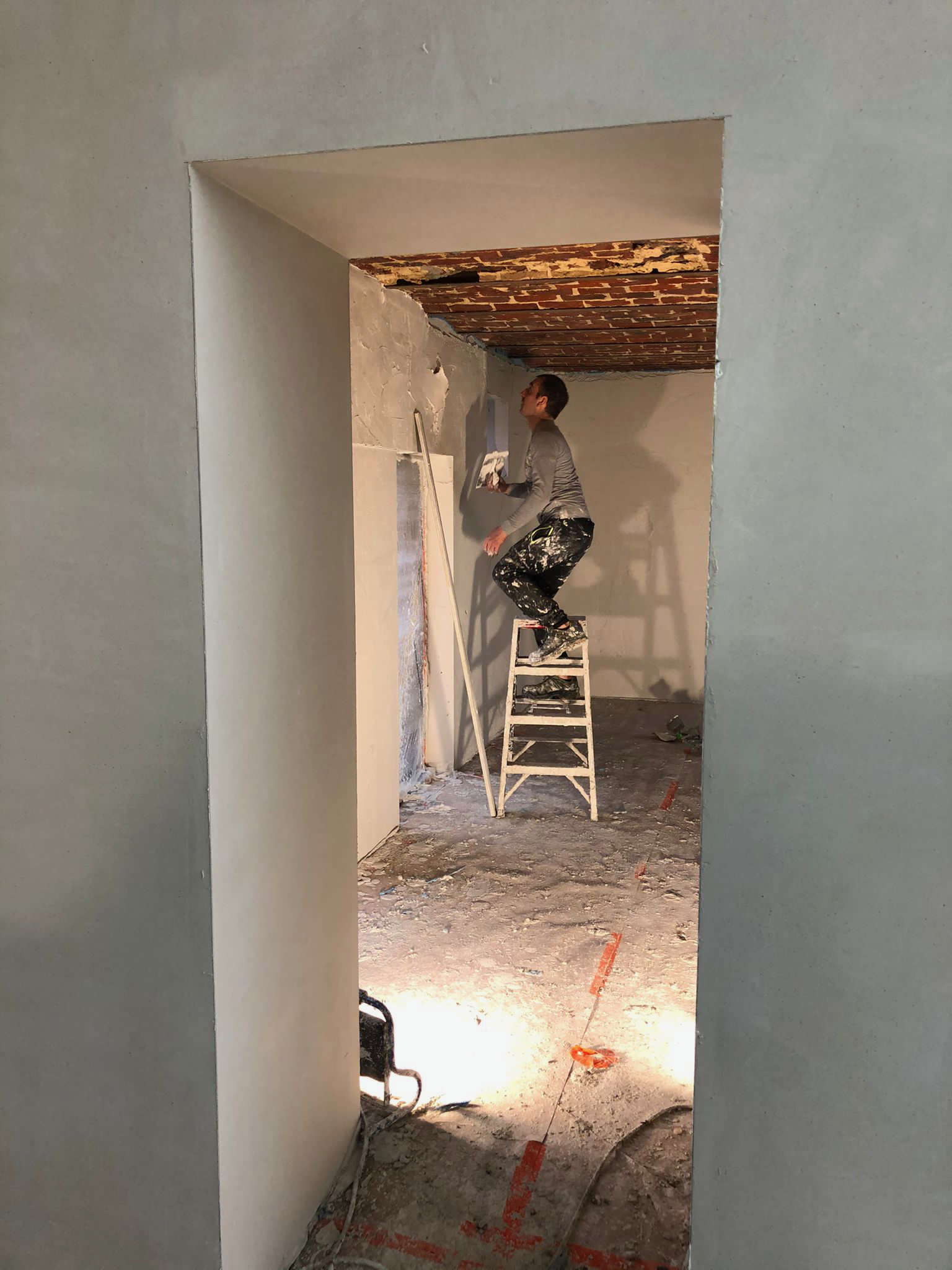 A person standing on a ladder while painting a wall in a room under renovation, with exposed brick ceiling and construction materials on the floor.
