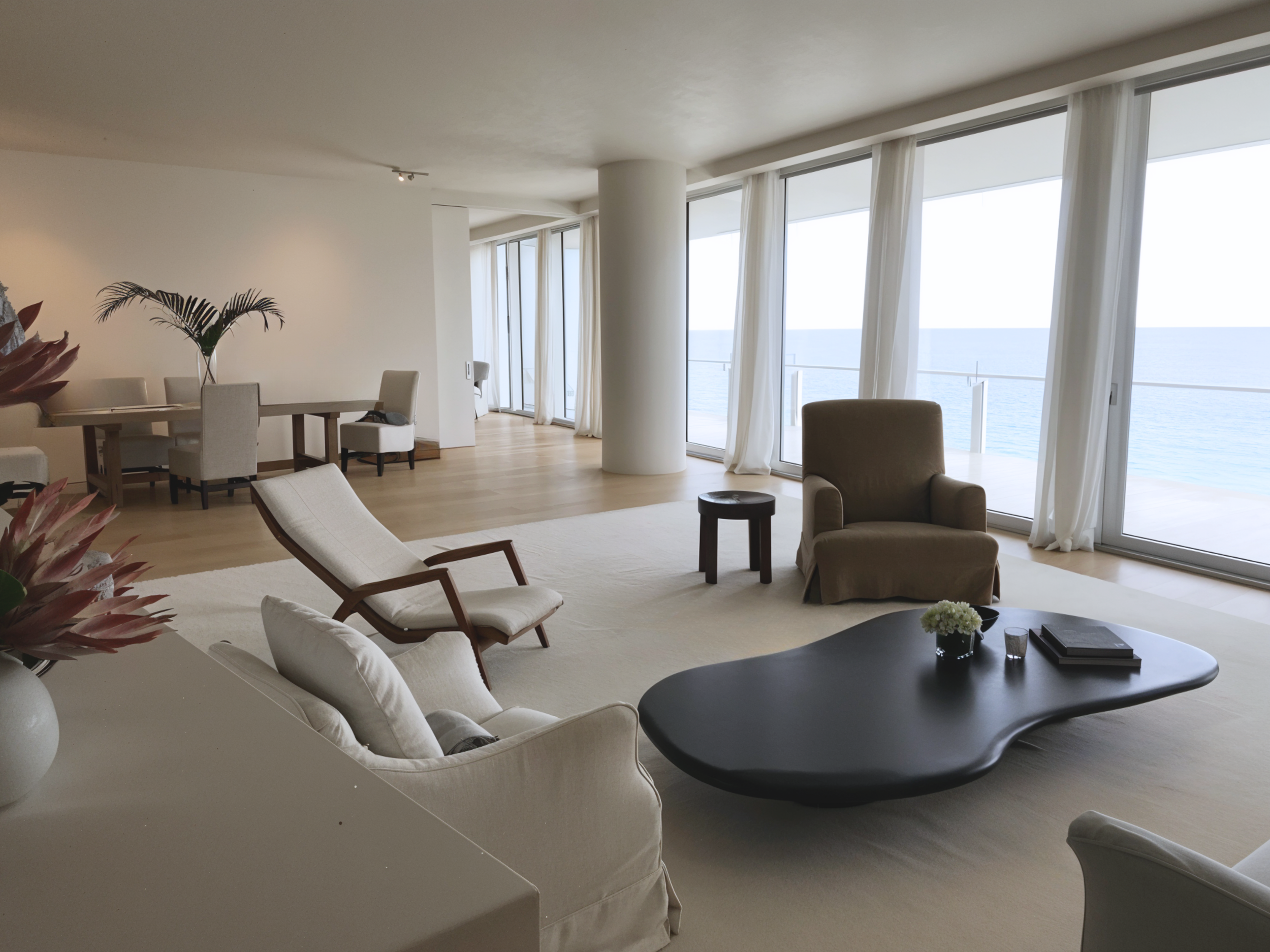JD Residence in Miami | Spacious modern living room with ocean view, featuring large windows, minimalist furniture, plush armchairs, a sleek black slatecoffee table, dining area with table and chairs, light wooden floors, and tropical decor elements.