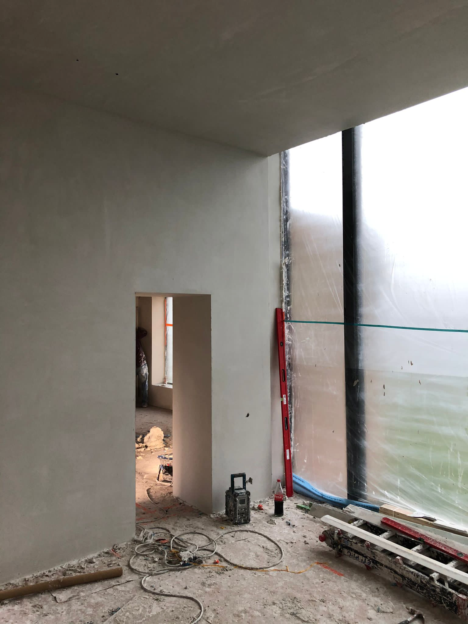 Construction site interior with unfinished plastered walls, a doorway, plastic-covered windows, and scattered construction tools and materials such as cables and a ladder.