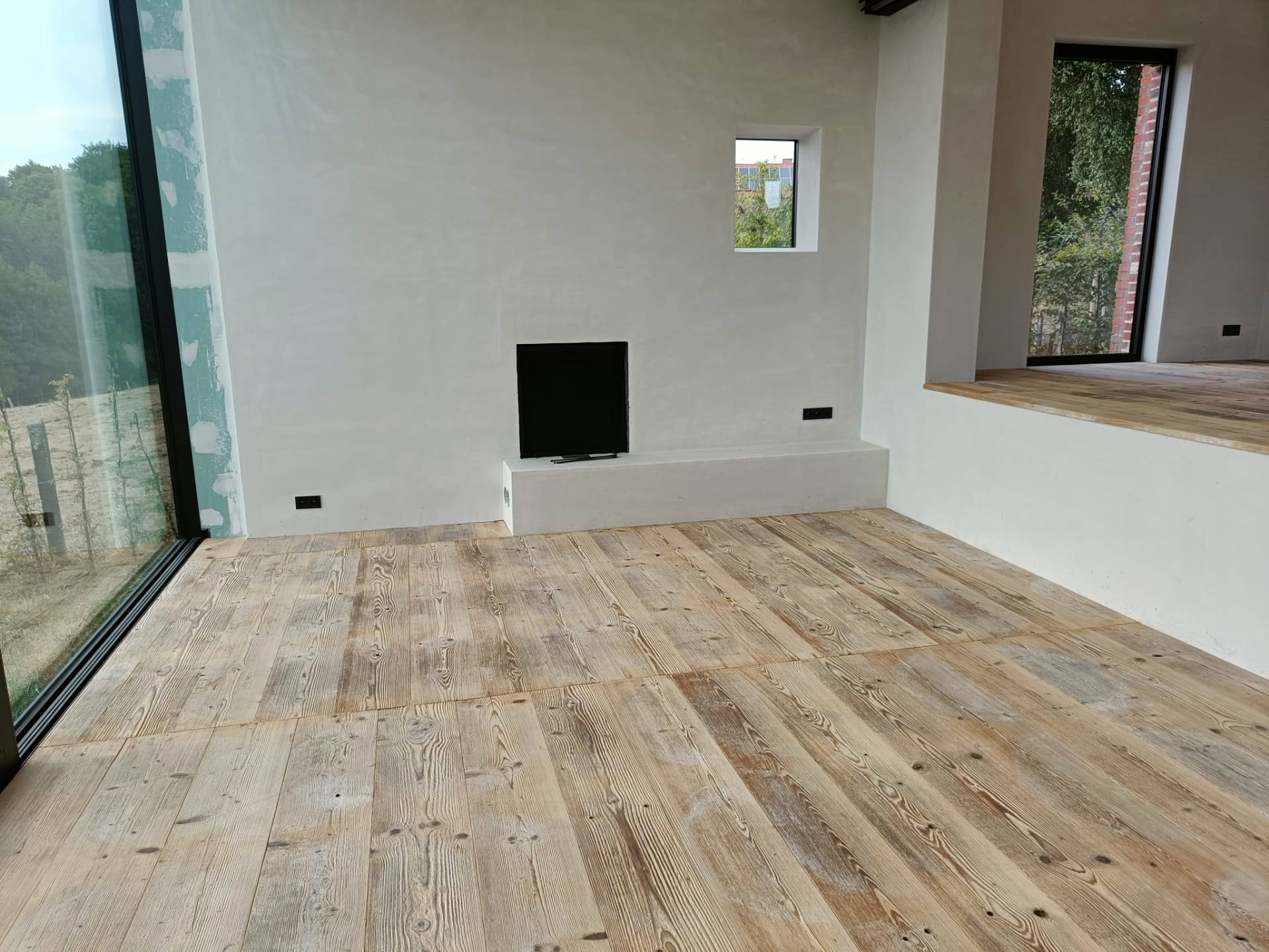 Modern minimalist room with natural wooden flooring, large glass windows, and plastered walls, featuring an open fireplace to the likings of Aires Mateus.
