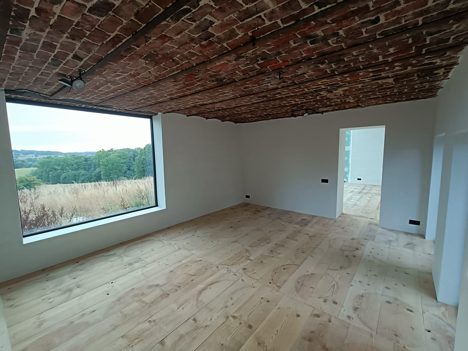 Empty room with brick ceiling, large window, and wooden floor overlooking a scenic landscape.