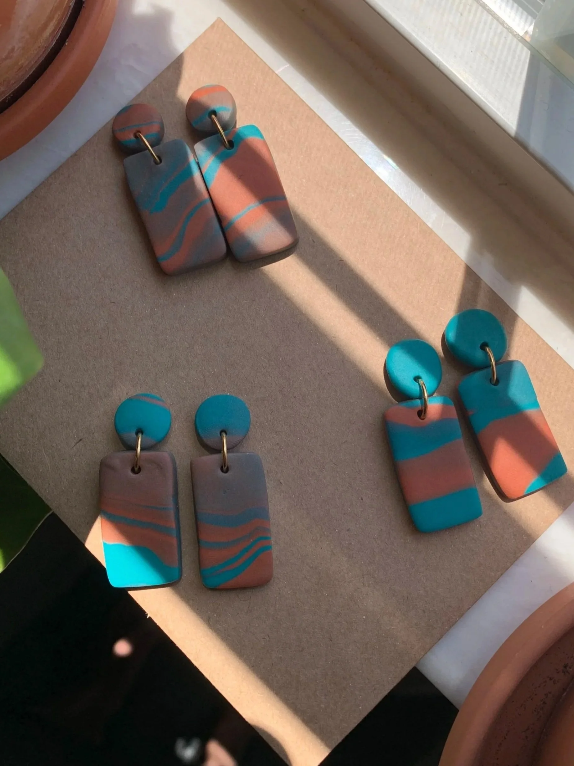 Three pairs of handmade dangle earrings with colorful marbled patterns in blue, pink, black, and beige, placed on a beige surface near a window with sunlight.