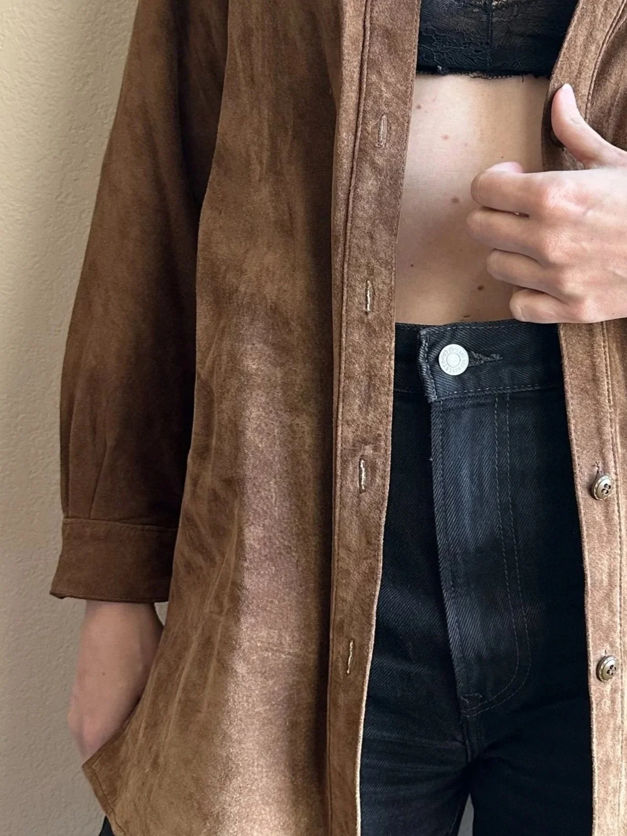Person wearing a brown suede jacket over a black top and black high-waisted jeans, with one hand adjusting the jacket.