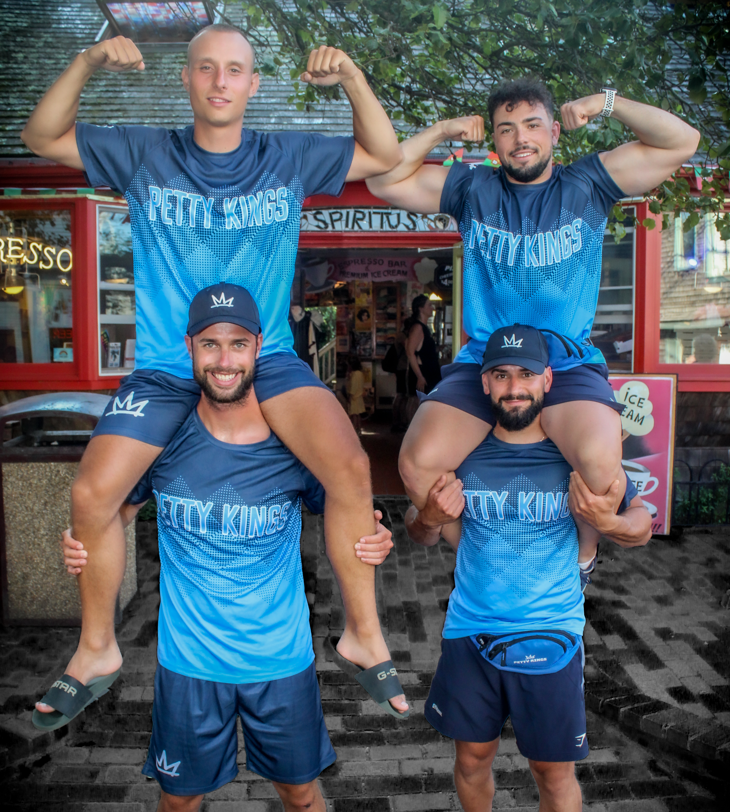 Two men wearing blue t-shirts with 'Petty Kings' written on them, flexing their biceps and smiling outdoors.