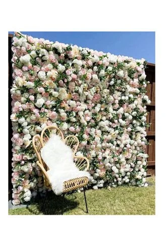 A chair with white faux fur draped over it, positioned in front of a large, lush floral wall made of pink and white roses, outdoors under a clear blue sky.