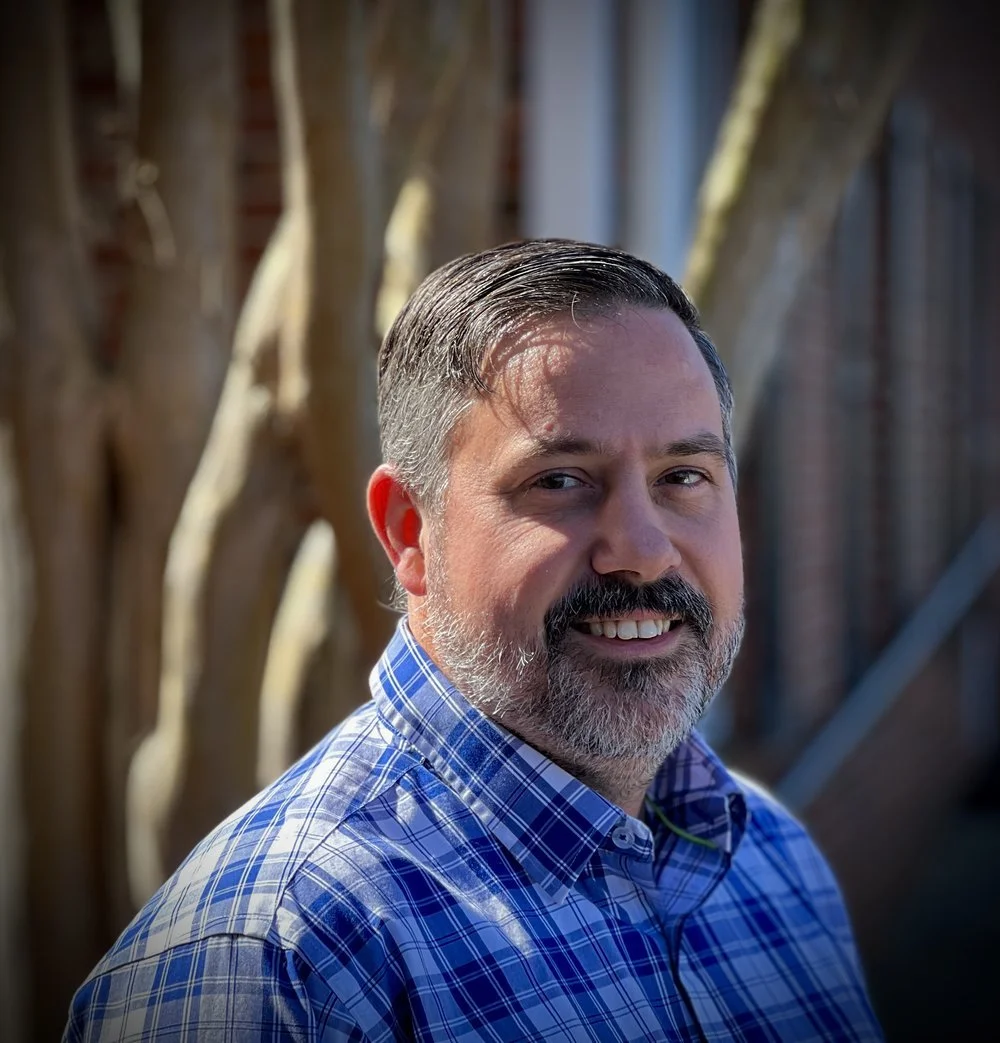 A man with short gray hair and a beard, wearing a blue plaid shirt, smiling outdoors with trees and a brick wall in the background.