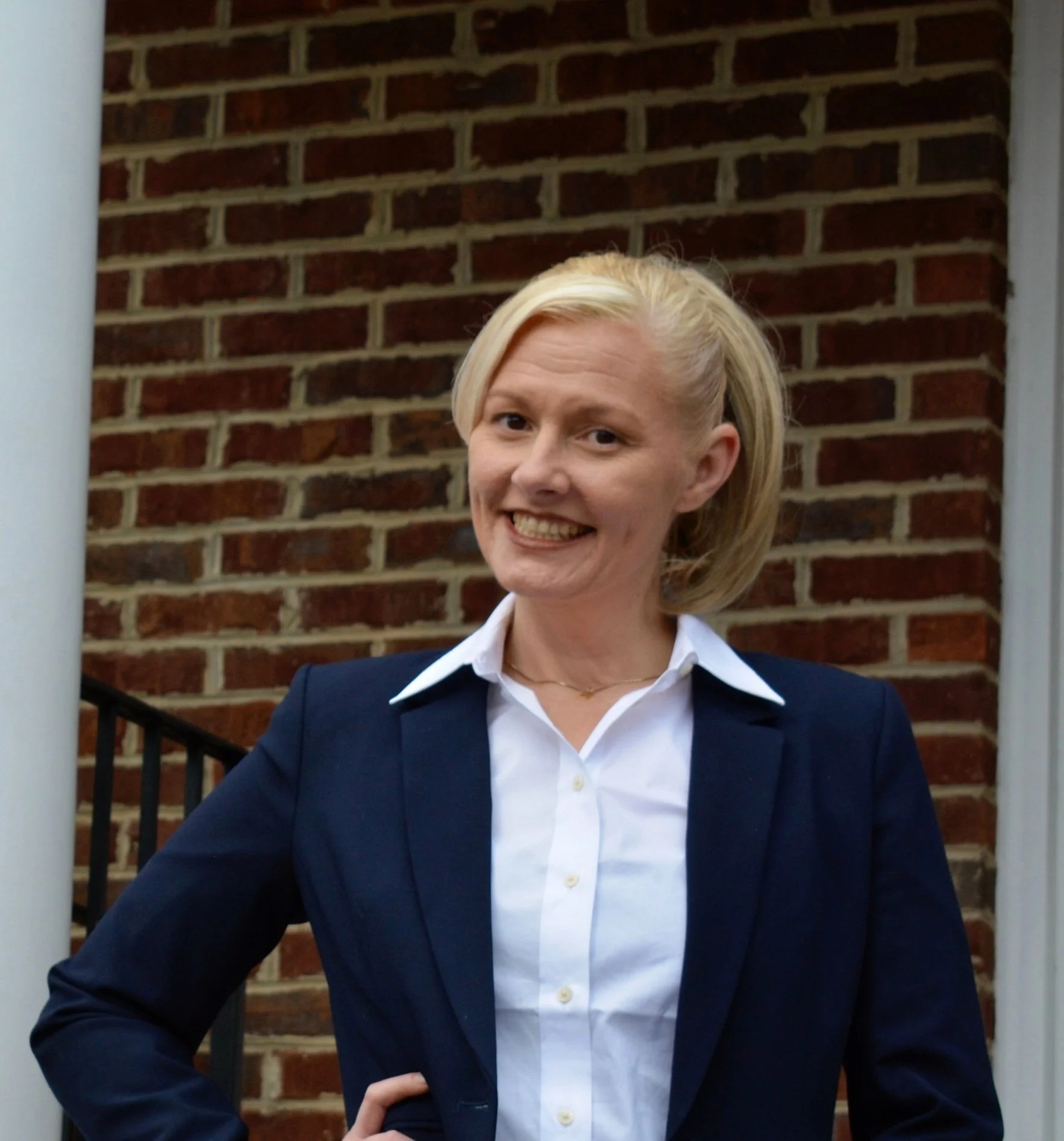 A woman with blonde hair dressed in a white shirt and navy blazer standing outdoors in front of a brick wall.