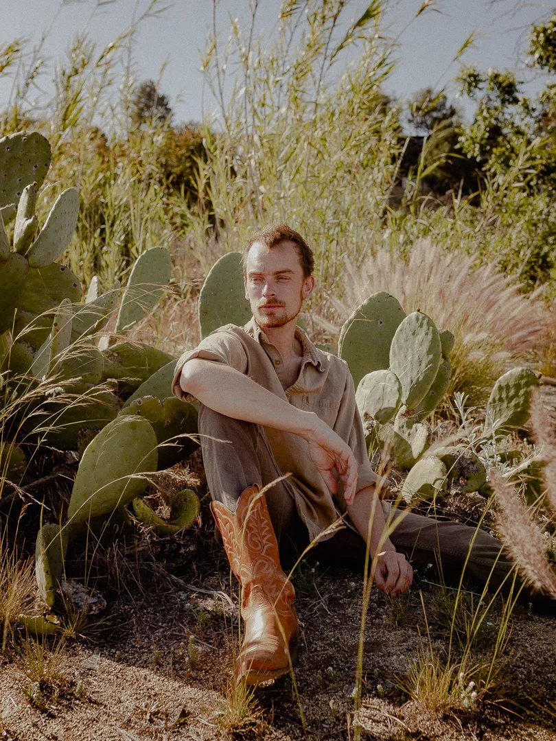 A man sitting among cactus plants and tall grasses in a desert landscape, wearing a beige shirt, dark pants, and cowboy boots, with sunlight casting shadows on his face.