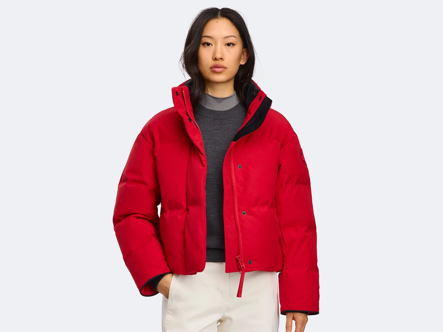 Young woman in a red puffer jacket, black and gray layered sweaters, and beige pants, standing against a plain white background.