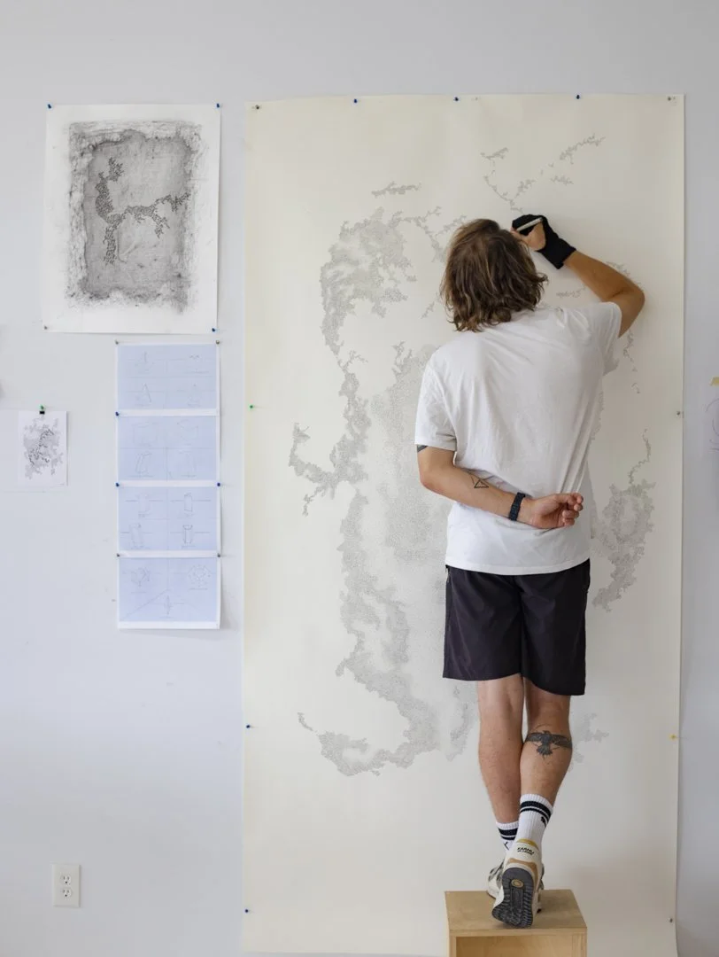 A person with brown hair, wearing a white T-shirt, black shorts, and sneakers, is standing on a wooden step stool and creating a large map of South America on a light-colored wall using a black marker.