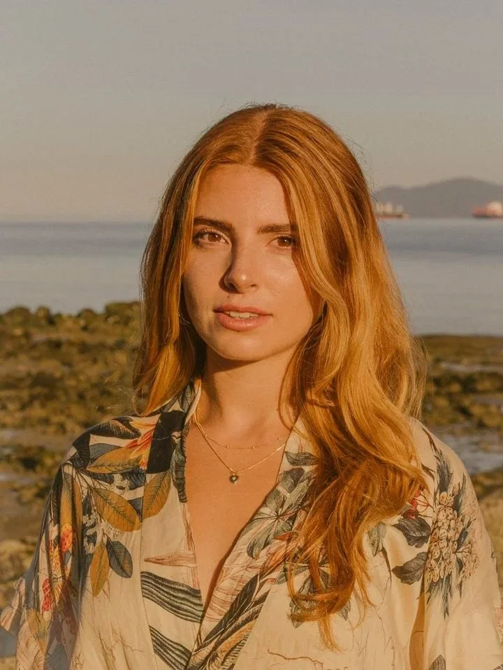 A woman with long red hair wearing a botanical patterned shirt, standing outdoors near water during sunset.