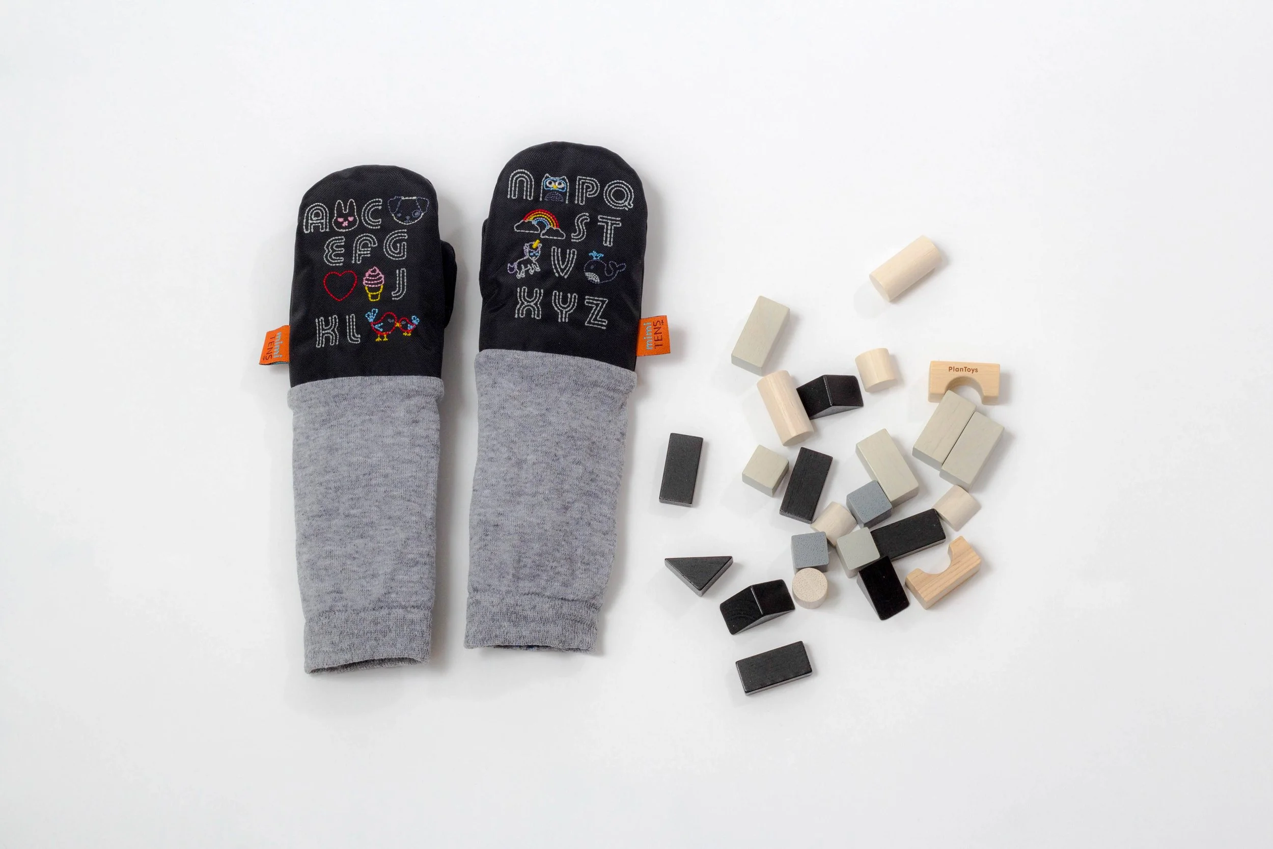 A pair of children's mittens with alphabet and animal embroidery on a white surface, next to scattered wooden blocks in black, gray, and natural wood tones.