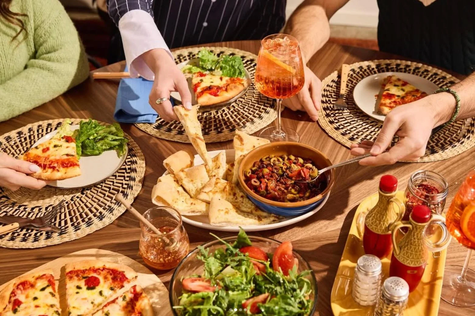 People sharing a meal at a wooden table with slices of pizza, a bowl of salad, a bowl of ragout or stew, and drinks, including a glass of rosé wine and a cocktail.
