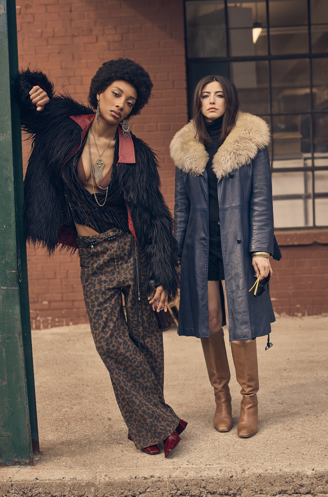 Two women standing outside near a brick building. One woman with an afro hairstyle wearing leopard print pants, a black and fur jacket, and red shoes. The other woman with shoulder-length brown hair wearing a navy blue coat with a large fur collar and brown boots.