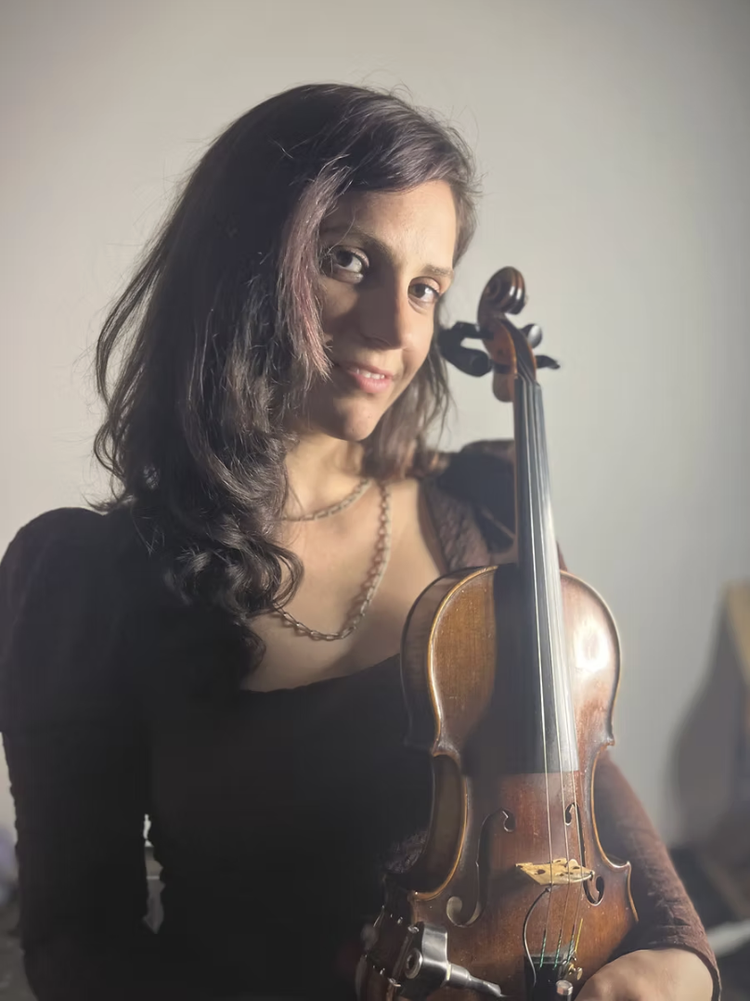 A woman with dark wavy hair holding a violin and smiling at the camera.