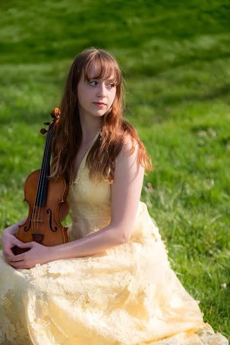 A young woman with long red hair sitting outdoors on grass, holding a violin in her lap, wearing a yellow dress.