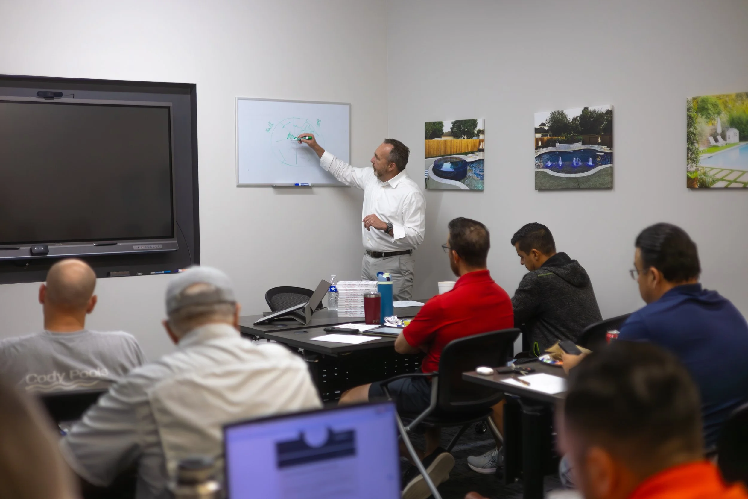 A man standing and speaking into a microphone in front of an audience in a conference room with beige walls. A presentation slide behind him reads 'Matt Martin' with a logo for 'Charge 20 San Antonio.' The audience is seated at tables with notebooks, laptops, and drinks, facing the speaker.