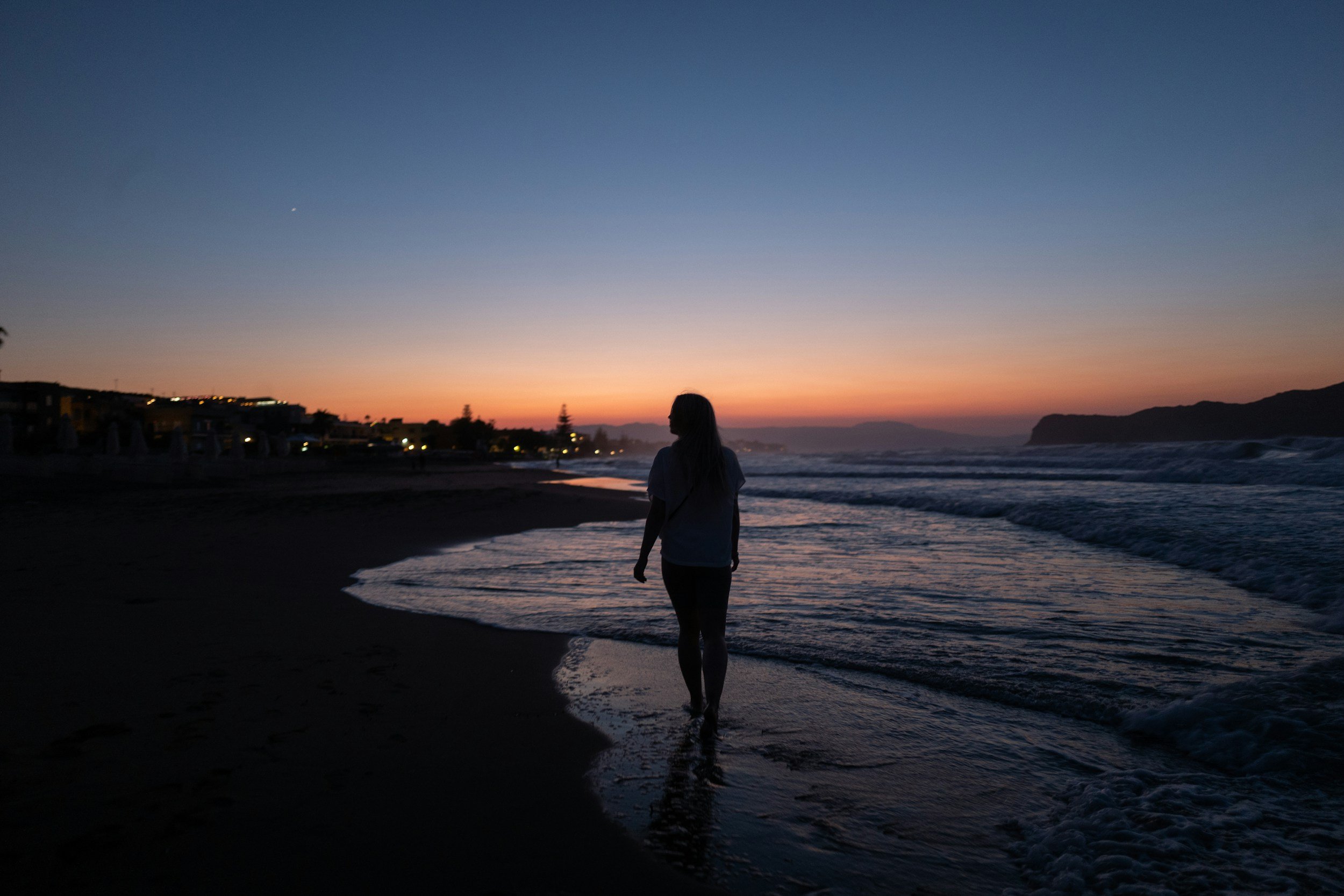 A woman walks along the beach at sunset with the sky transitioning from orange near the horizon to deep blue. There are buildings with lights on along the shoreline and mountains in the distance.