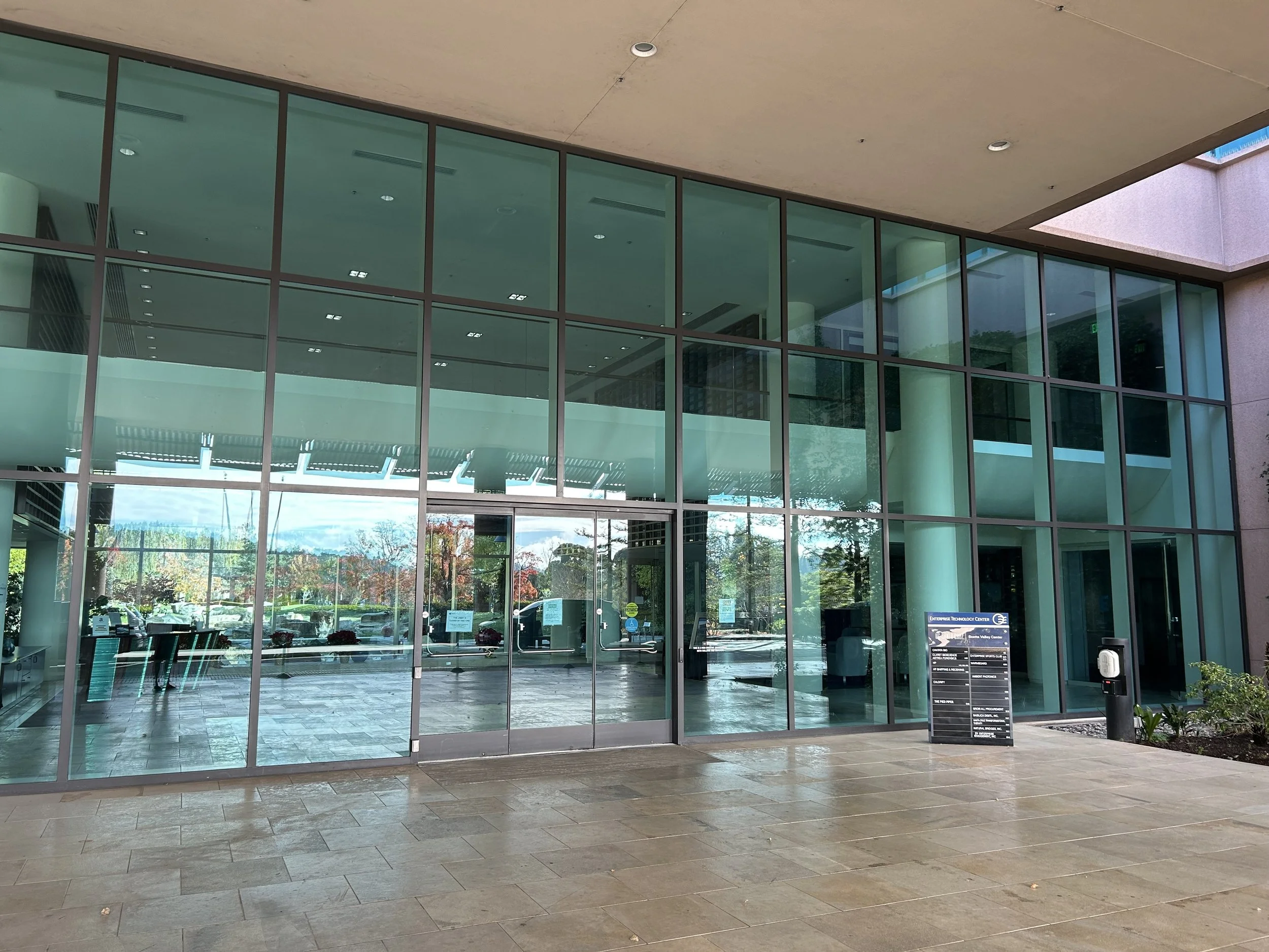 Modern glass entrance to a building with reflection of trees and cars outside, and a black directory sign on the right.