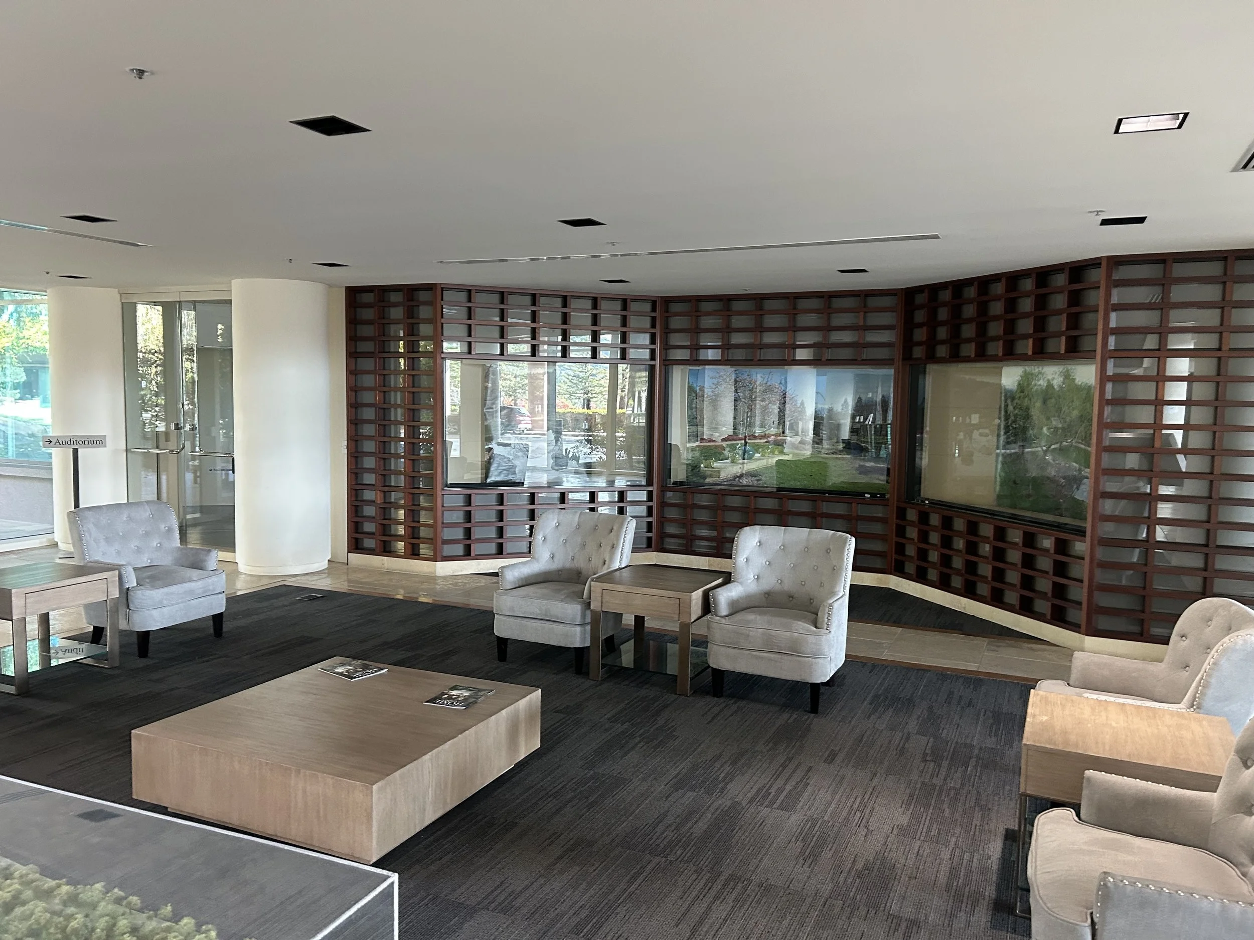 A modern interior lobby with beige armchairs, a wooden coffee table, dark carpet, large windows, and a wooden lattice structure on the wall.