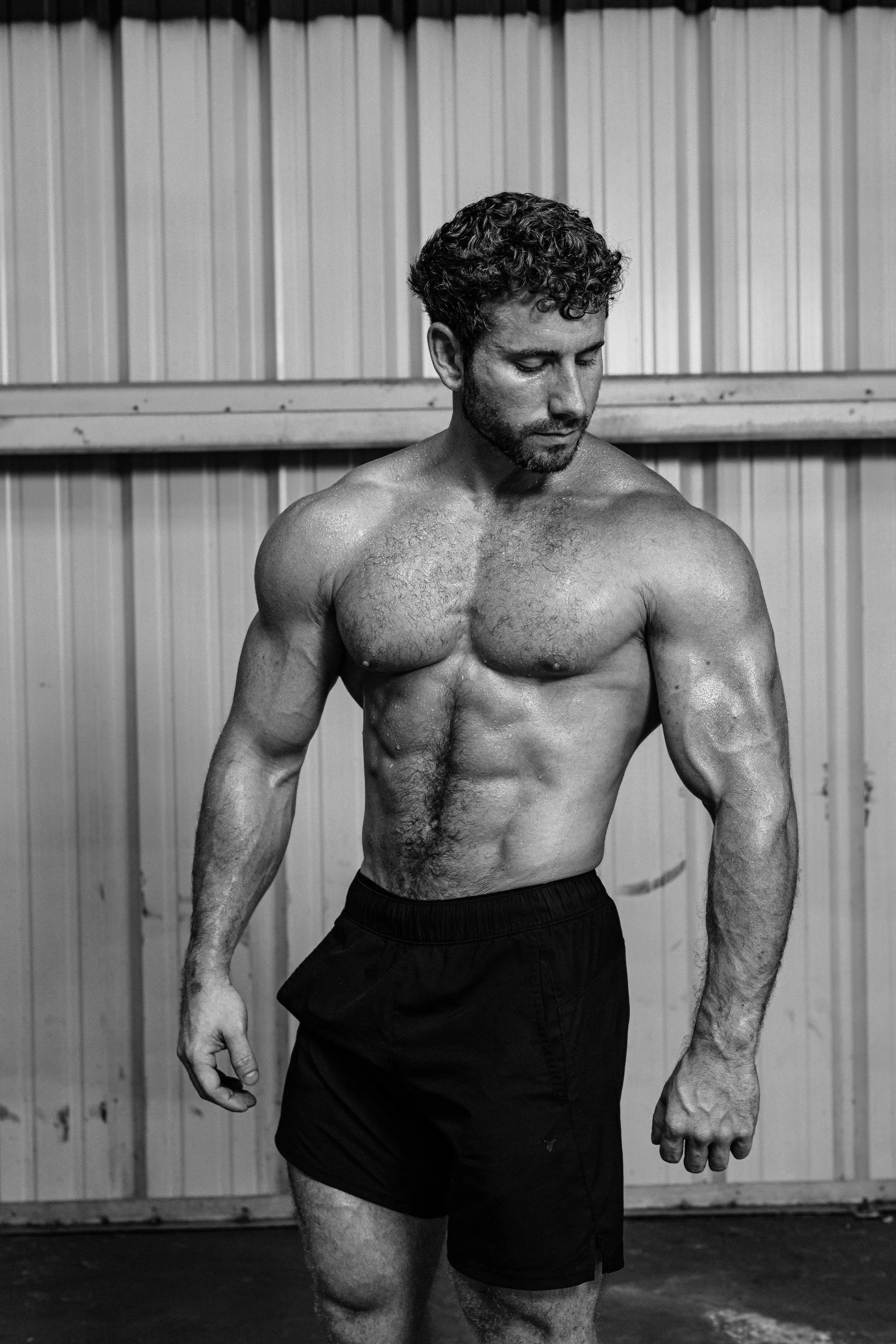 A shirtless man with curly hair and a beard, wearing black shorts, is standing in front of a wooden wall, looking downward.