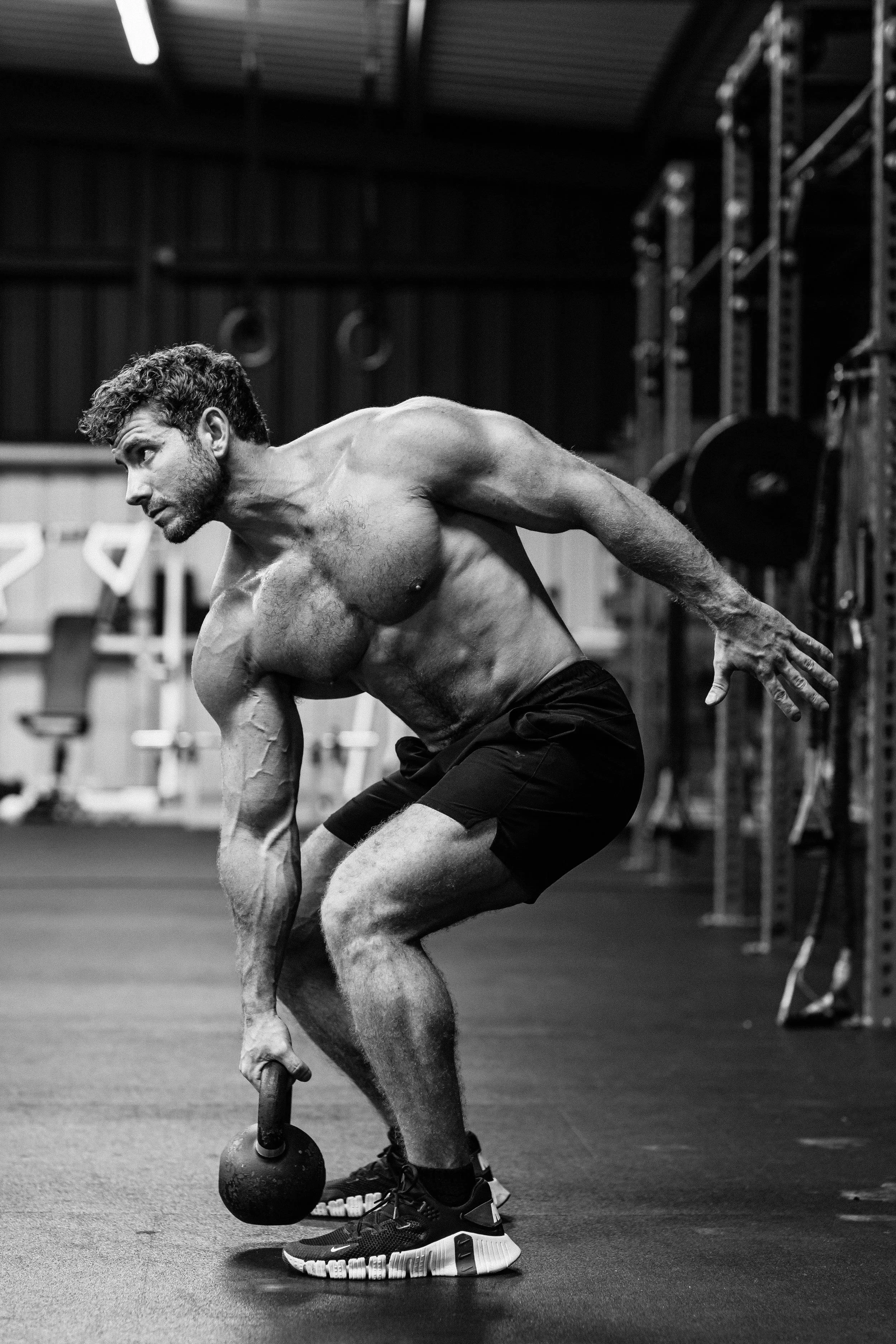 A shirtless man with curly hair exercising with a kettlebell in a gym. He is in a bent-over position, lifting the kettlebell with one hand while balancing on one foot. Equipment, shelves, and gym walls are visible in the background.