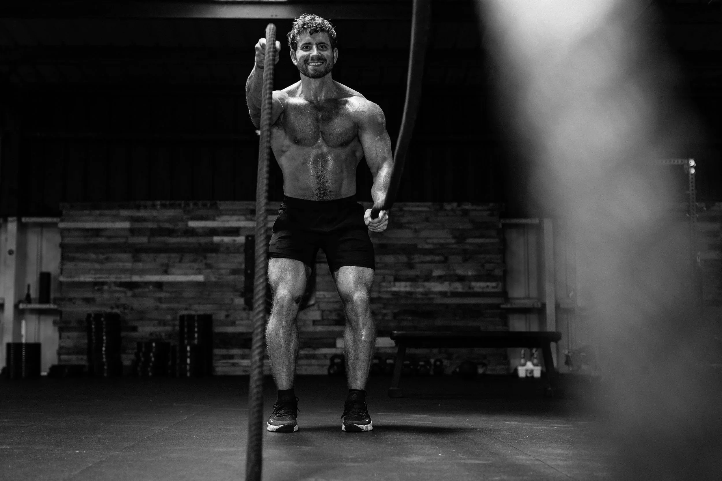 A shirtless man with a beard and curly hair smiling while holding a rope in a gym setting with wooden wall panels, weightlifting equipment, and gym mats.
