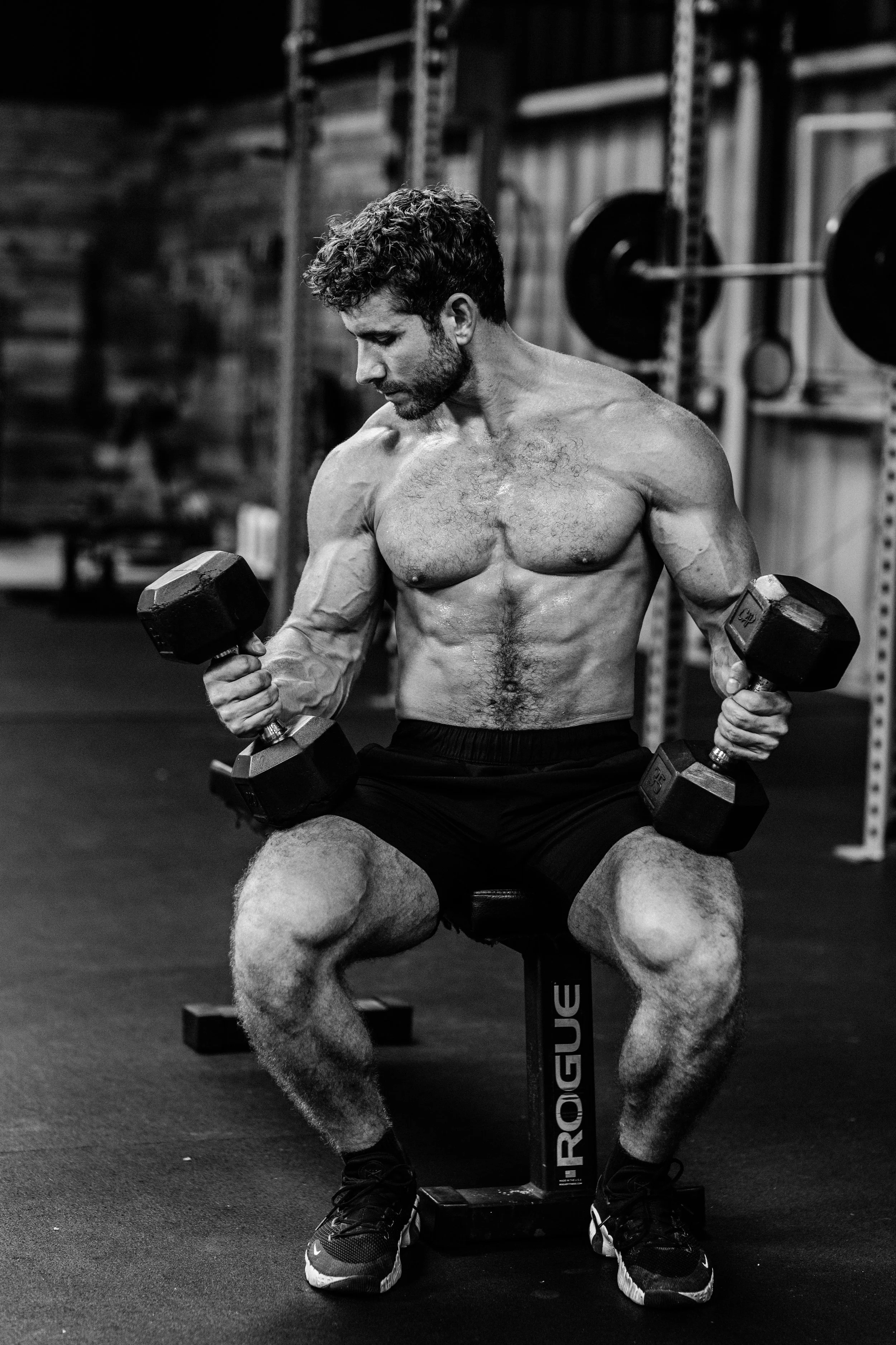 A shirtless man with curly hair and a beard sitting on a stool in a gym, lifting dumbbells, with a focused expression. The gym has weight racks in the background.
