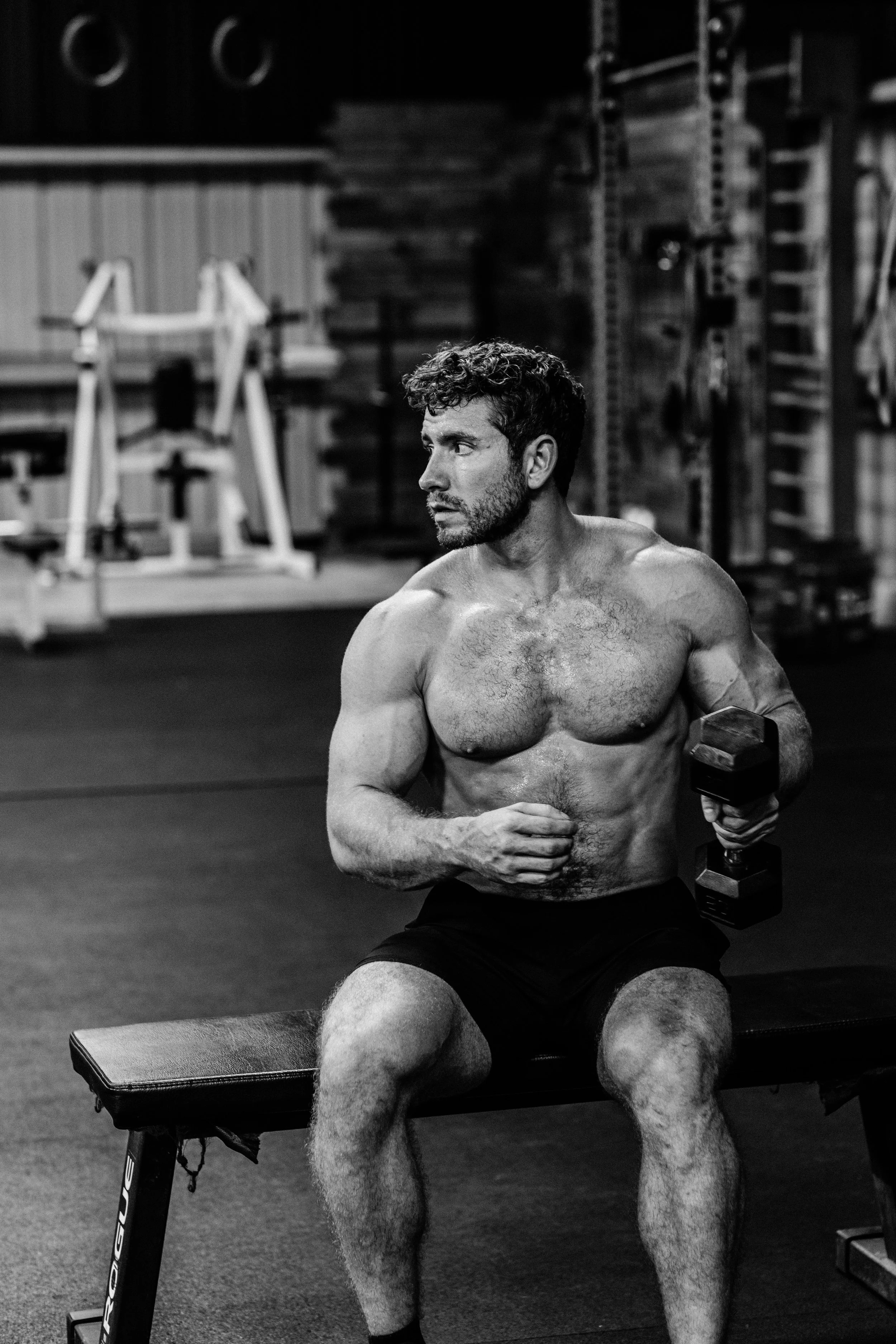 A shirtless man with curly hair and a beard working out in a gym, sitting on a bench and holding a dumbbell in his right hand.