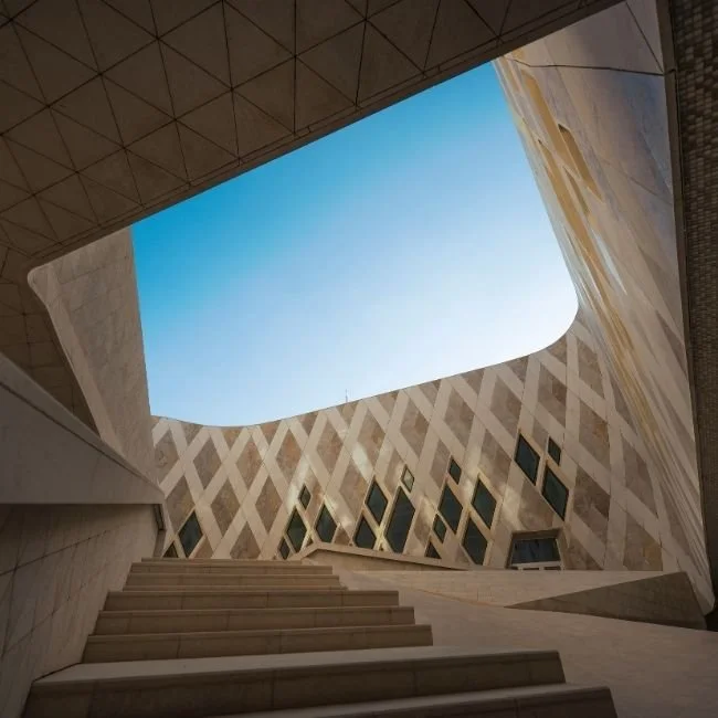 Vue de l'intérieur d'un bâtiment moderne avec un ciel visible à travers une ouverture au plafond, murs en pierre avec des fenêtres en forme de losange, et un escalier en pierre.