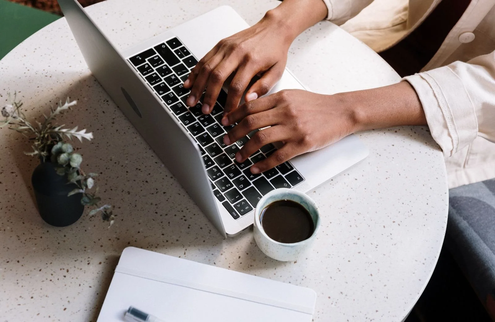 Personne tapant sur un clavier d'ordinateur portable blanc, assise devant une table ronde avec une tasse de café, un carnet blanc et un petit vase avec des plantes sèches.