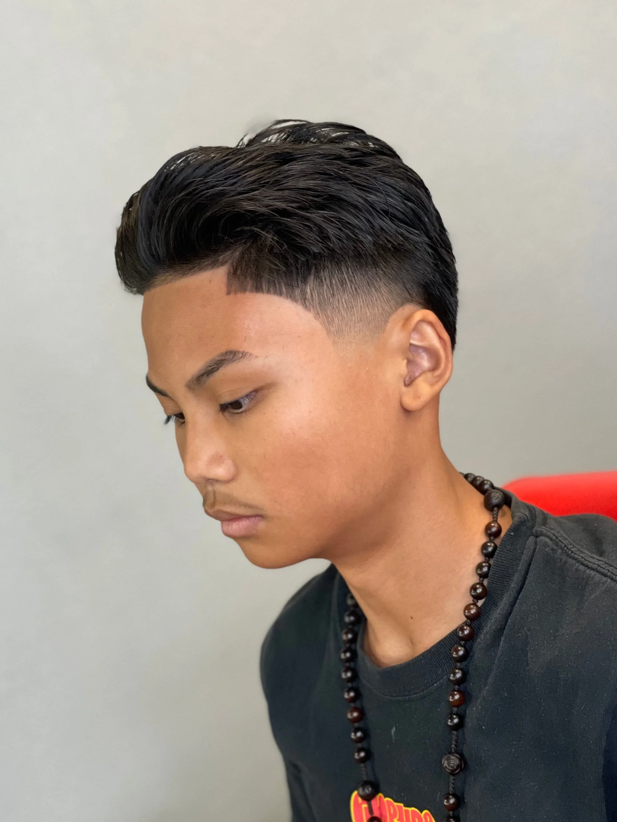 Side profile of a young man with a modern, slick hairstyle featuring a fade and a comb-over by Yoboy the Barber - Las Vegas barbershop. He is wearing a black shirt and a beaded necklace, sitting against a plain gray wall.