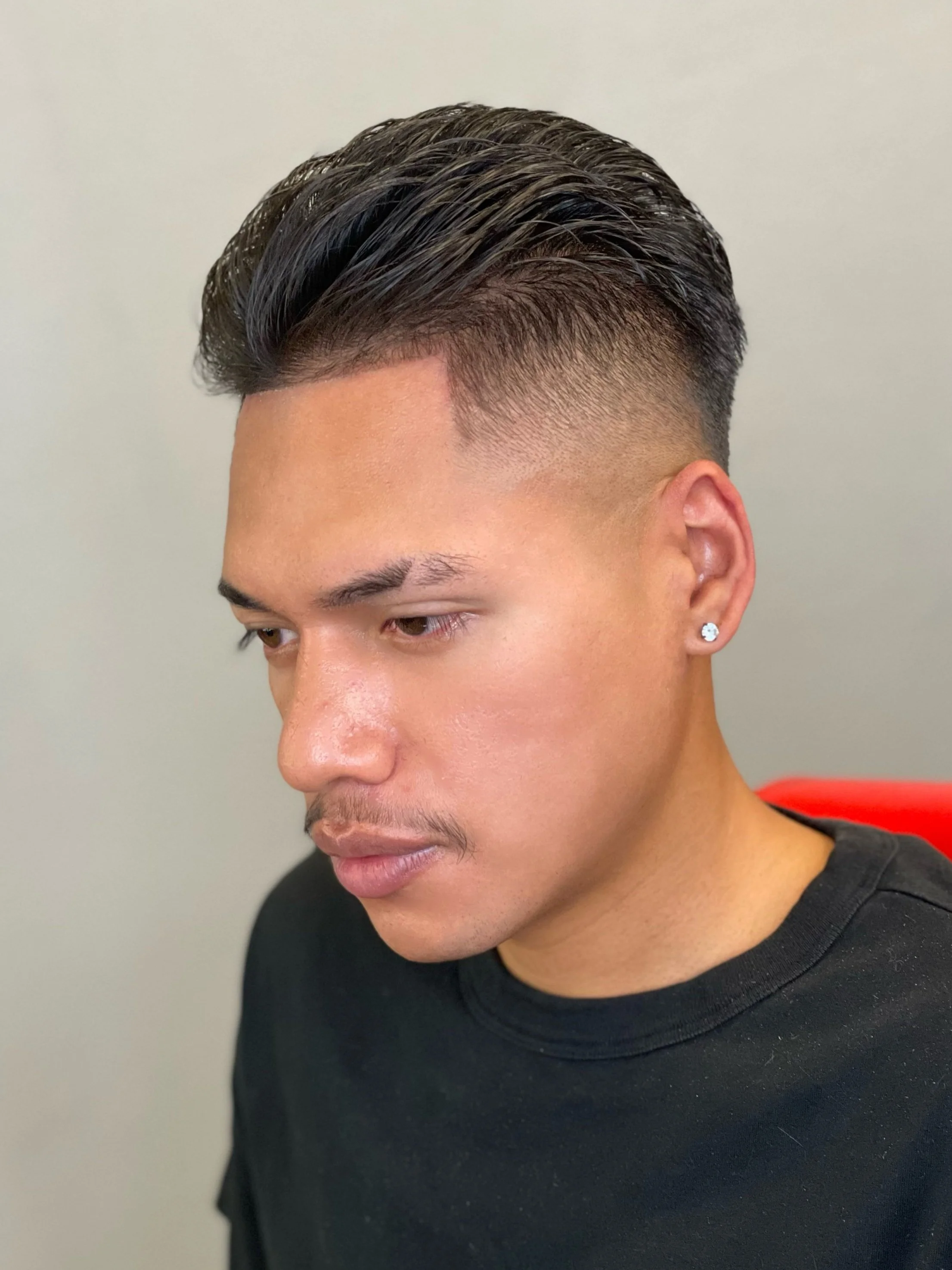 Young man with a modern fade haircut and styled brushback haircut by Yoboy the Barber - Las Vegas barber. He is wearing a diamond earring, black t-shirt, looking slightly downward, neutral background.