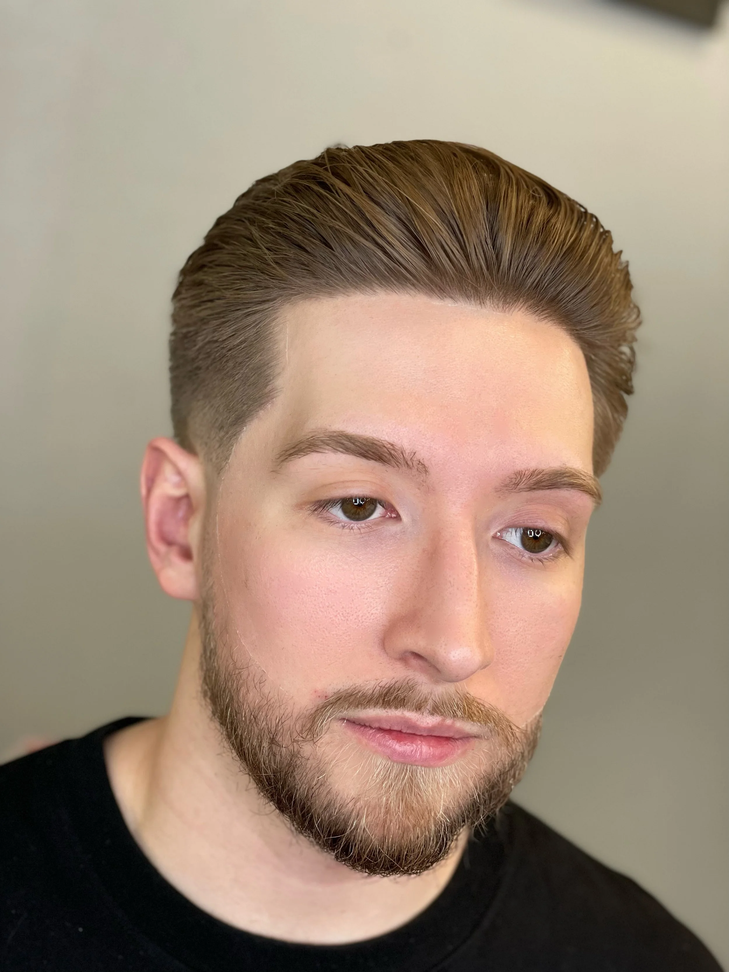 Close-up of a young man with styled brown hair slick back and low drop fade haircut, lined up beard, and groomed eyebrows by Yoboy the Barber - Las Vegas barbershop. He has brown eyes, and a beard, wearing a black shirt, looking slightly down.
