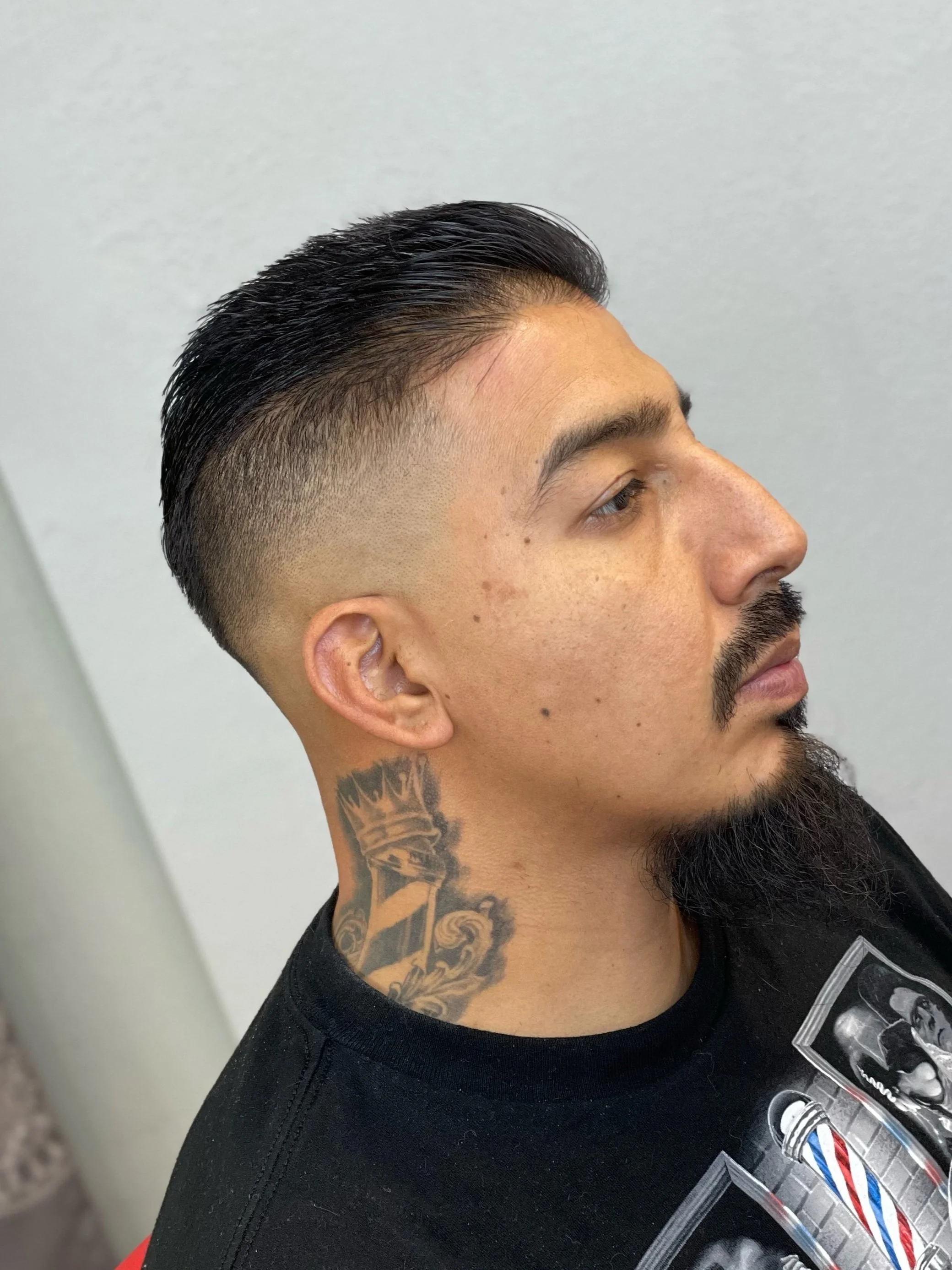 Side profile of a man with a fresh undercut hairstyle, dark slicked-back hair, haircut by Yoboy the Barber - Las Vegas barbershop. There is a tattoo of a crown on his neck, wearing a black shirt with graphic print.
