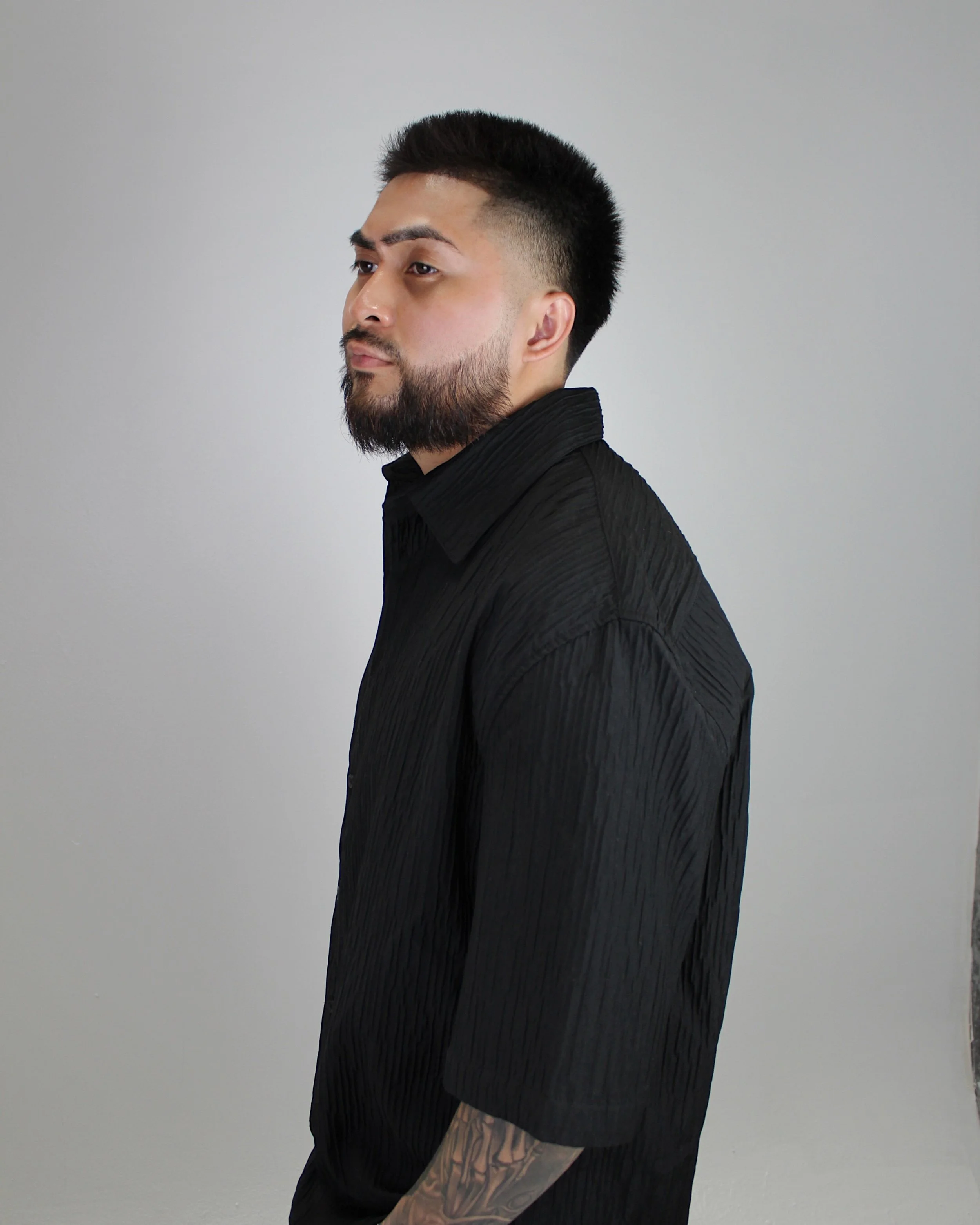 Side profile of a man with a beard and tattoos wearing a black textured shirt against a plain light gray background. Yoboy the Barber - best barber in Las Vegas