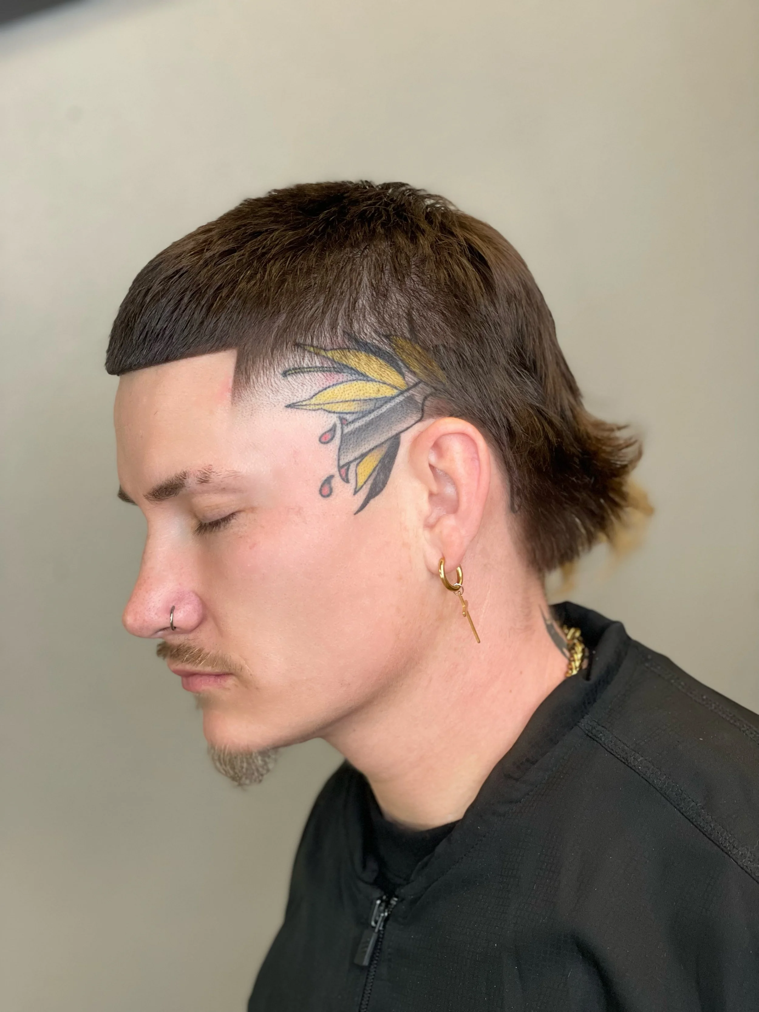 Close-up side profile of a man with brown hair, tattoos on his face and ear, wearing hoop earrings and a black jacket, with his eyes closed. High taper haircut, mullet, sharp lineup, and eyebrows done by Yoboy the Barber - Las Vegas barbershop.