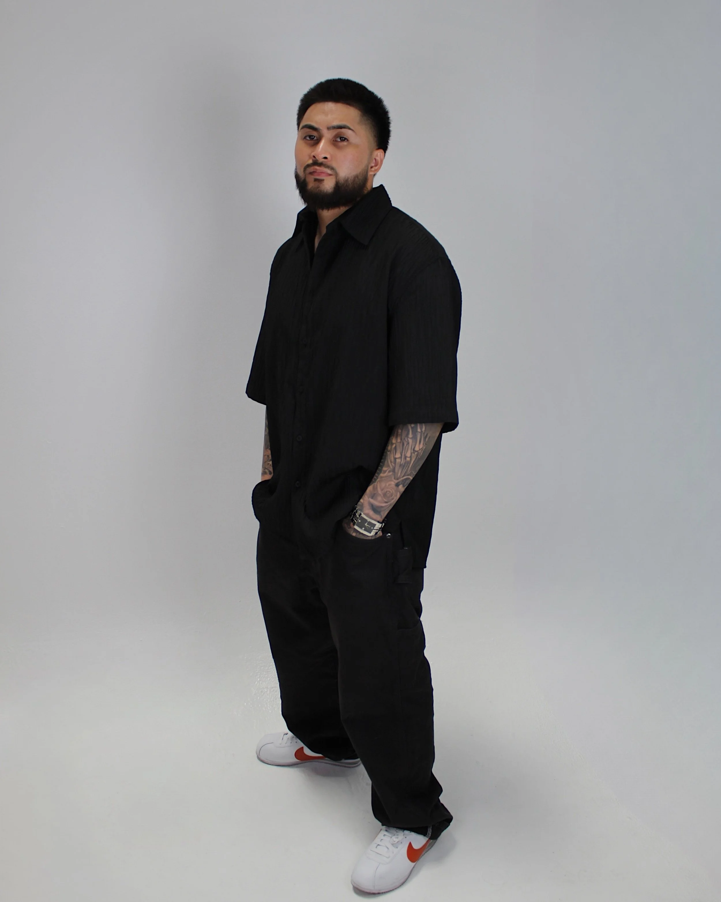 Yoboy the Barber - Best Las Vegas Mobile Barber. A man with dark hair, beard, and tattoos on his arm, wearing a black shirt, black pants, white sneakers with orange swoosh, standing with hands in his pockets against a plain light gray background.