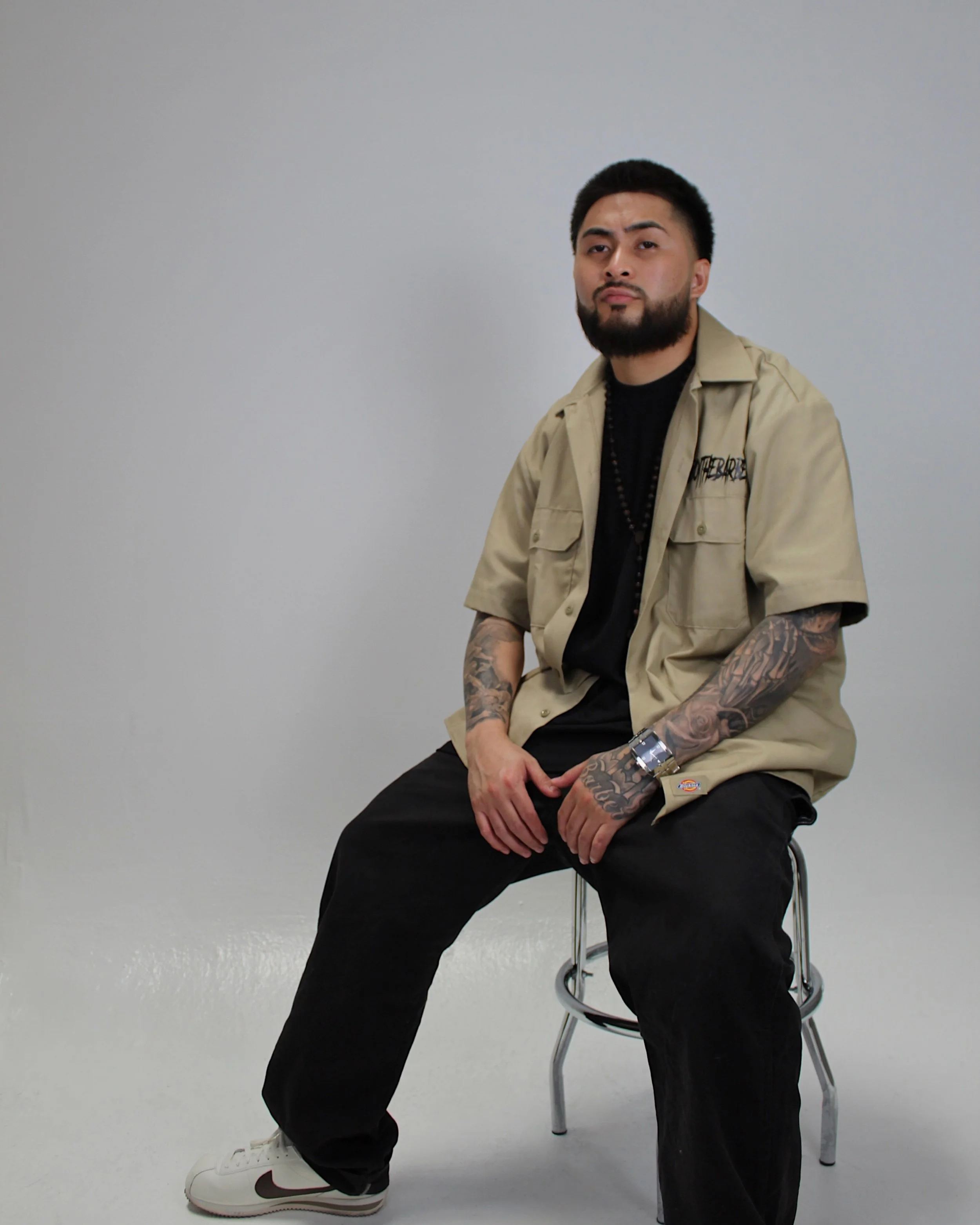 A man with a beard and tattoos on his arms sitting on a stool in a plain white room, wearing a beige short-sleeve shirt, black pants, white sneakers, and a black t-shirt underneath. Yoboy the Barber - Las Vegas Mobile Barber.