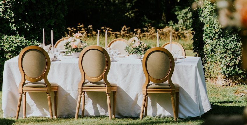 Une grande table dressée à l'extérieur, avec une nappe blanche, des chaises beige, et des décorations florales, probablement préparée pour un événement ou un repas.