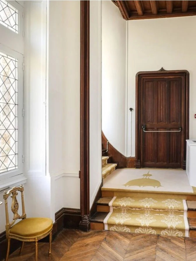 Entrée avec escaliers, tapis avec pattern dauphin et chien, porte en bois, fenêtre avec grilles, chaise ancienne en bois et tissu jaune.