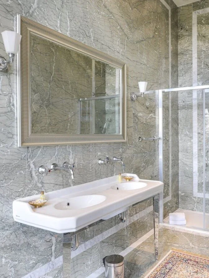 Salle de bain avec double lavabo blanc, grand miroir encadré, murs en marbre gris, cabine de douche avec porte en verre, tapis oriental, petit panier en verre avec produits dans la salle de bain.