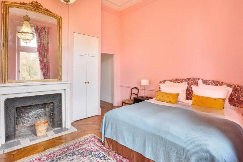 Chambre élégante avec mur rose, lit avec tête de lit en tissu à motif floral, deux coussins jaunes, une table de nuit avec lampe, cheminée en marbre, grand miroir doré, et tapis oriental.