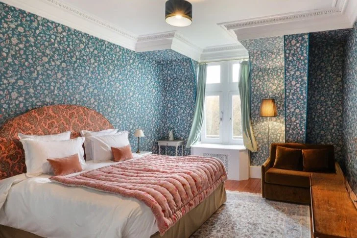 Chambre avec Mur à papier peint fleuri, grand lit avec oreillers blancs et coussins marron, lampes de chevet, fenêtre avec rideaux verts et bleus, fauteuil marron avec lampe de sol, tapis beige.