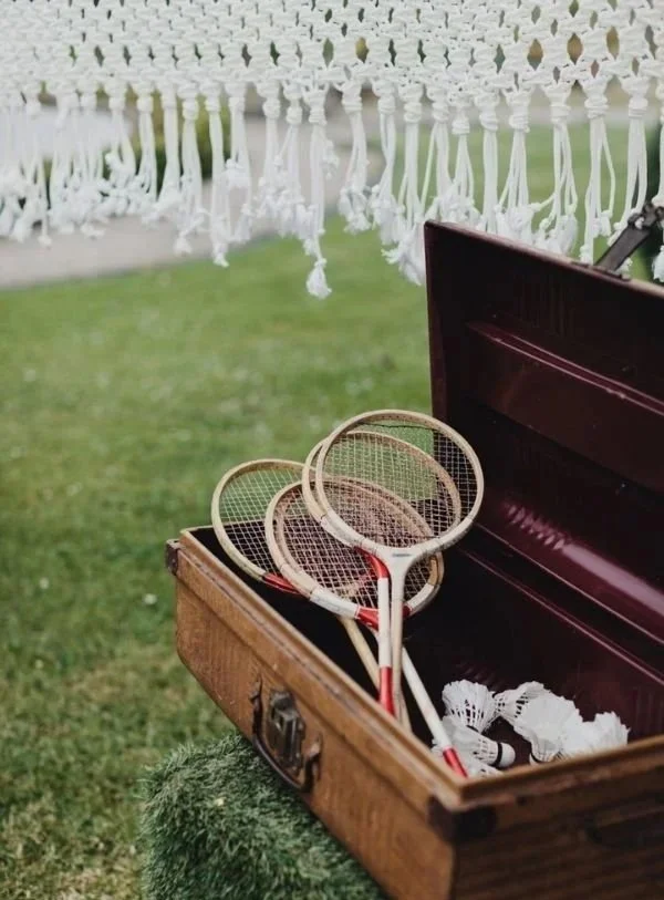 Un trousseau de tennis avec quatre raquettes à l'intérieur d'une vieille valise en bois ouverte, sur une pelouse, avec des plumes de badminton suspendues en haut.