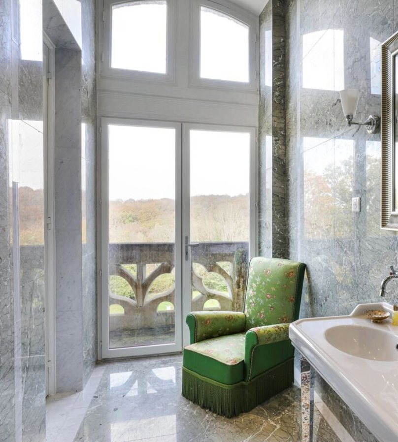 Une salle de bain avec un fauteuil vert à motifs floraux, une fenêtre donnant sur un balcon en pierre, et un lavabo blanc. Le mur est en marbre gris veiné.