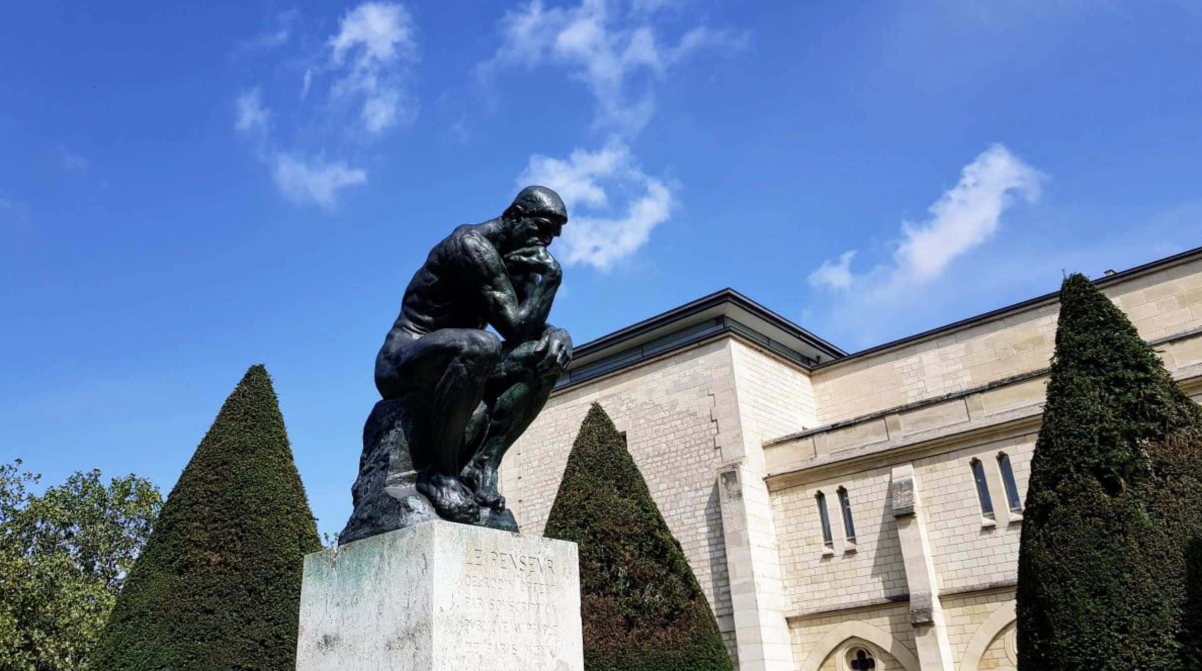 Statue de réflexion sur un socle en pierre, située dans un jardin avec des arbres taillés en forme de cône et un bâtiment en pierre en arrière-plan, sous un ciel bleu avec quelques nuages.